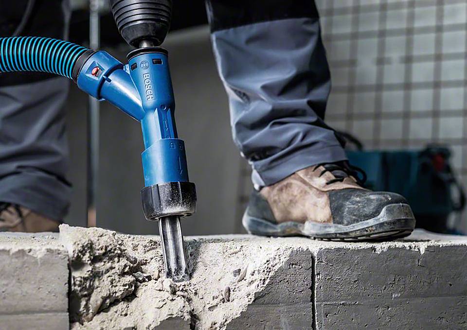 Een persoon boort met een elektrische boormachine en afzuiginstallatie in een bakstenen muur. Beschermende kleding en stevige schoenen zijn zichtbaar.