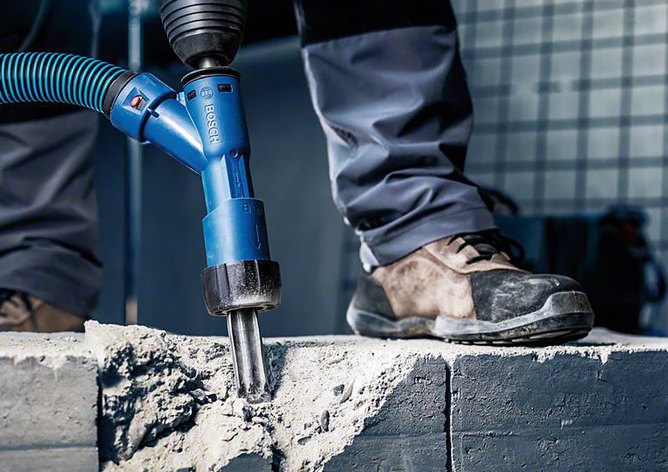 Een persoon boort in beton met een blauwe boorhamer, draagt veiligheidsschoenen; stofafzuiging is aangesloten.