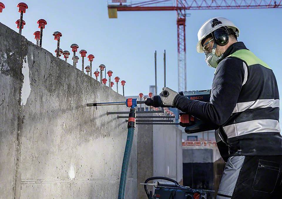 Een bouwvakker in veiligheidskleding boort een gat in een betonnen muur op een bouwterrein, met een kraan op de achtergrond.