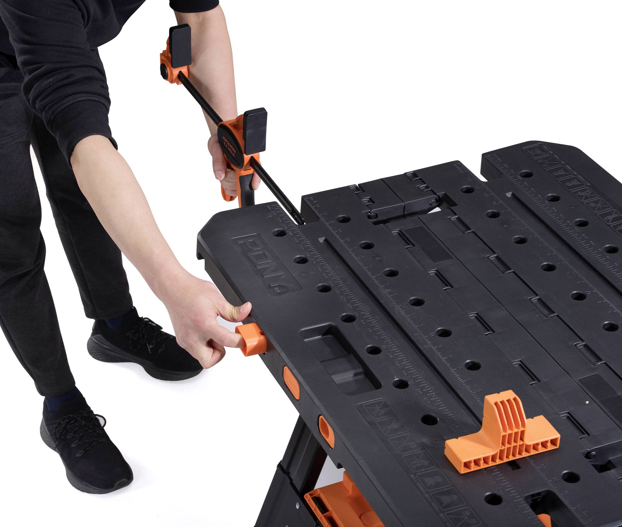 Een persoon gebruikt een bankschroef om een werkstuk op een werktafel vast te zetten. De tafel is zwart met oranje details.