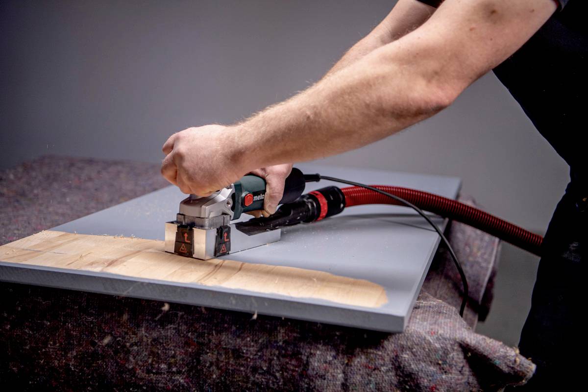 Een persoon bewerkt een houten plaat met een elektrisch gereedschap. Houtkrullen zijn zichtbaar terwijl het gereedschap een gladde oppervlakte creëert.