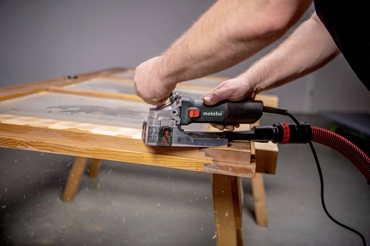Een persoon schuurt een houten tafel met een elektrische schuurmachine. Stofdeeltjes zijn zichtbaar.