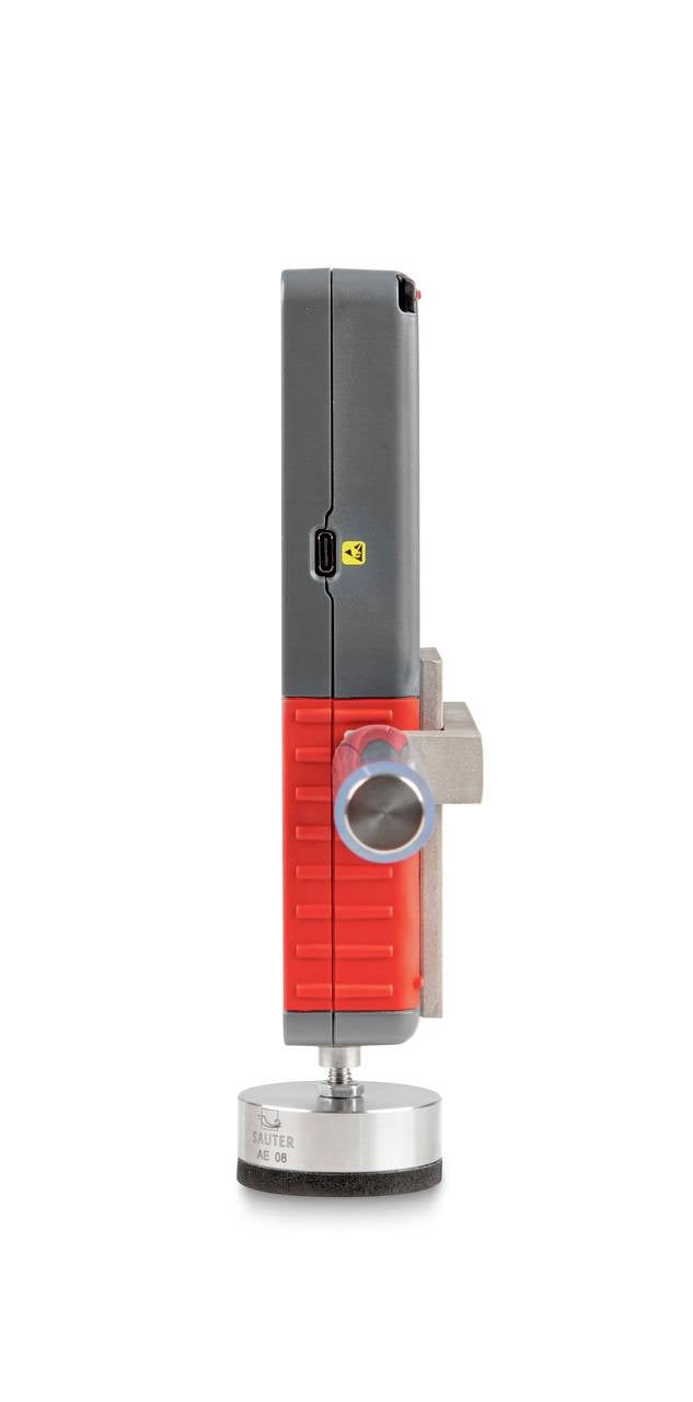 Een rood-zwart meetapparaat met een cilindrische basis en een zijknop, staat rechtop op een witte oppervlakte.