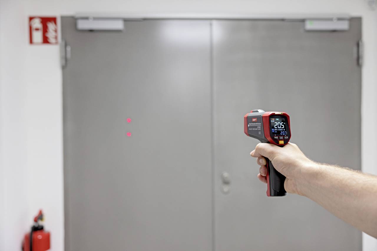 Een hand houdt een infrarood-thermometer vast, gericht op een grijze deur. Het display toont een temperatuur van 20,5 graden Celsius.