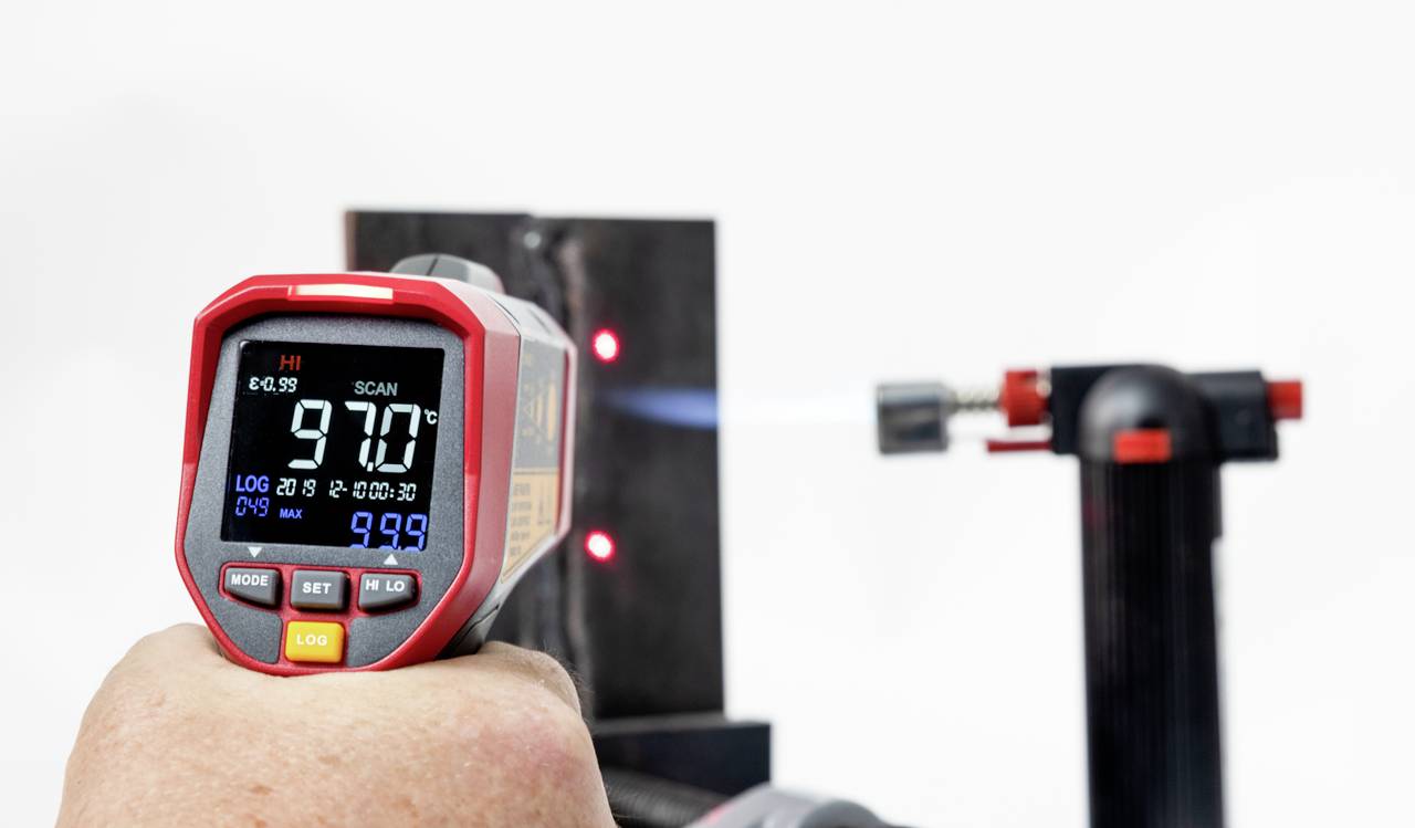Een hand houdt een infrarood-thermometer vast dat een temperatuur van 97,0 graden aanwijst. Op de achtergrond is een vlam zichtbaar.