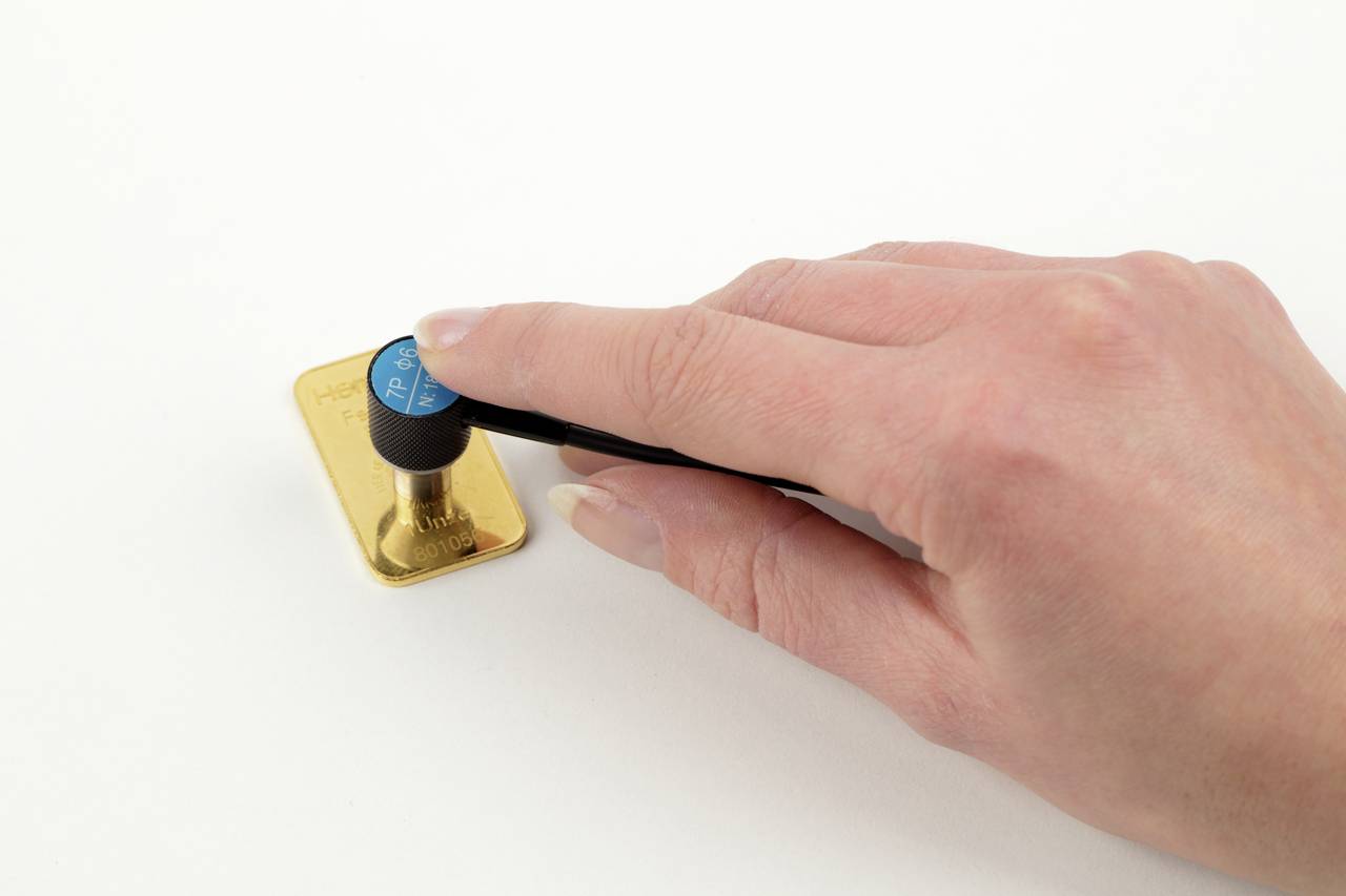 Een hand drukt met een zwart gereedschap een blauwe knop op een gouden plaat tegen een witte achtergrond.