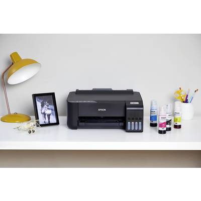 Een tafel met een printer in het midden. Links een lamp en een ingelijste foto, rechts inktflesjes en een pennenhouder.