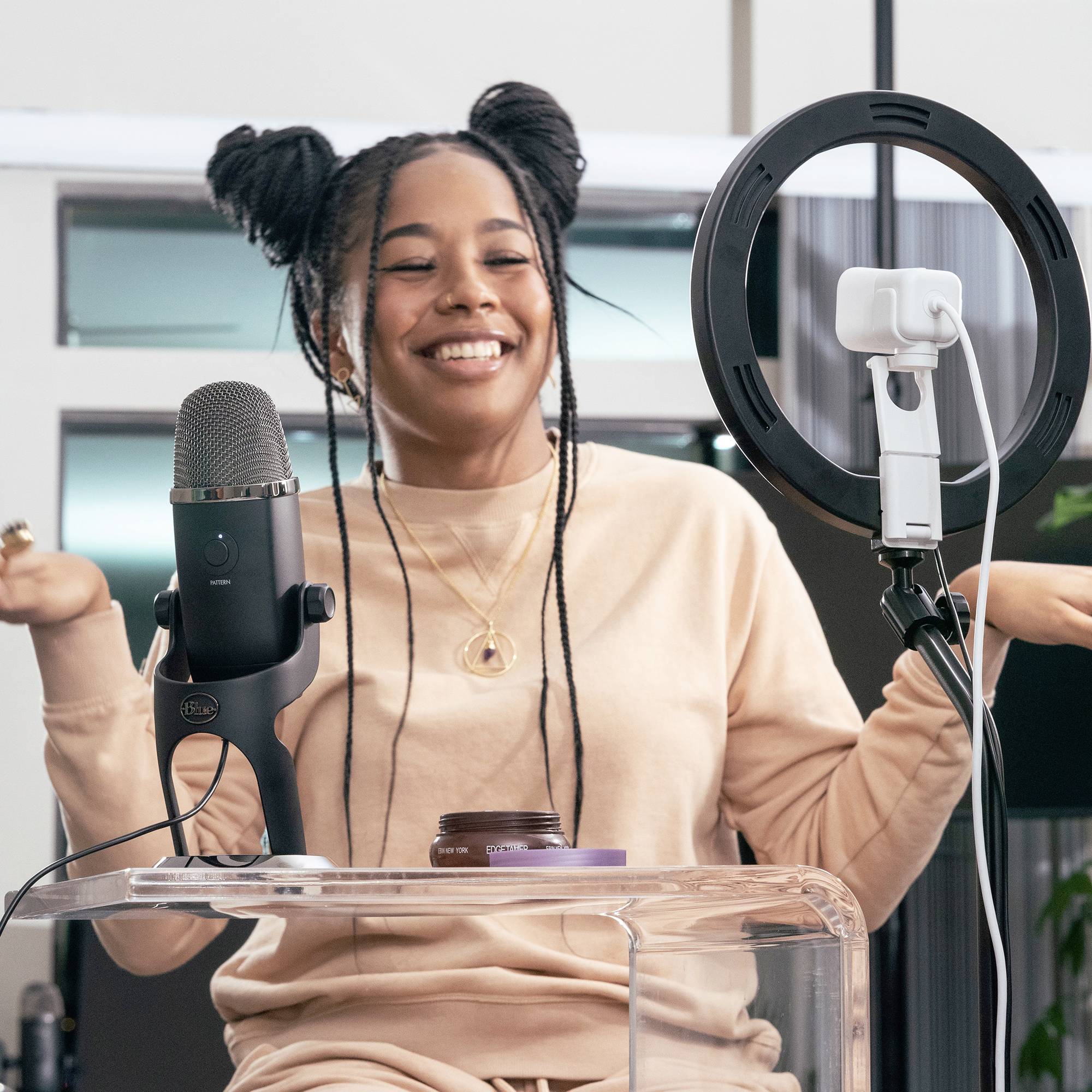 Een persoon zit glimlachend voor een microfoon en een ringlight, klaar om op te nemen of te streamen in een studio.