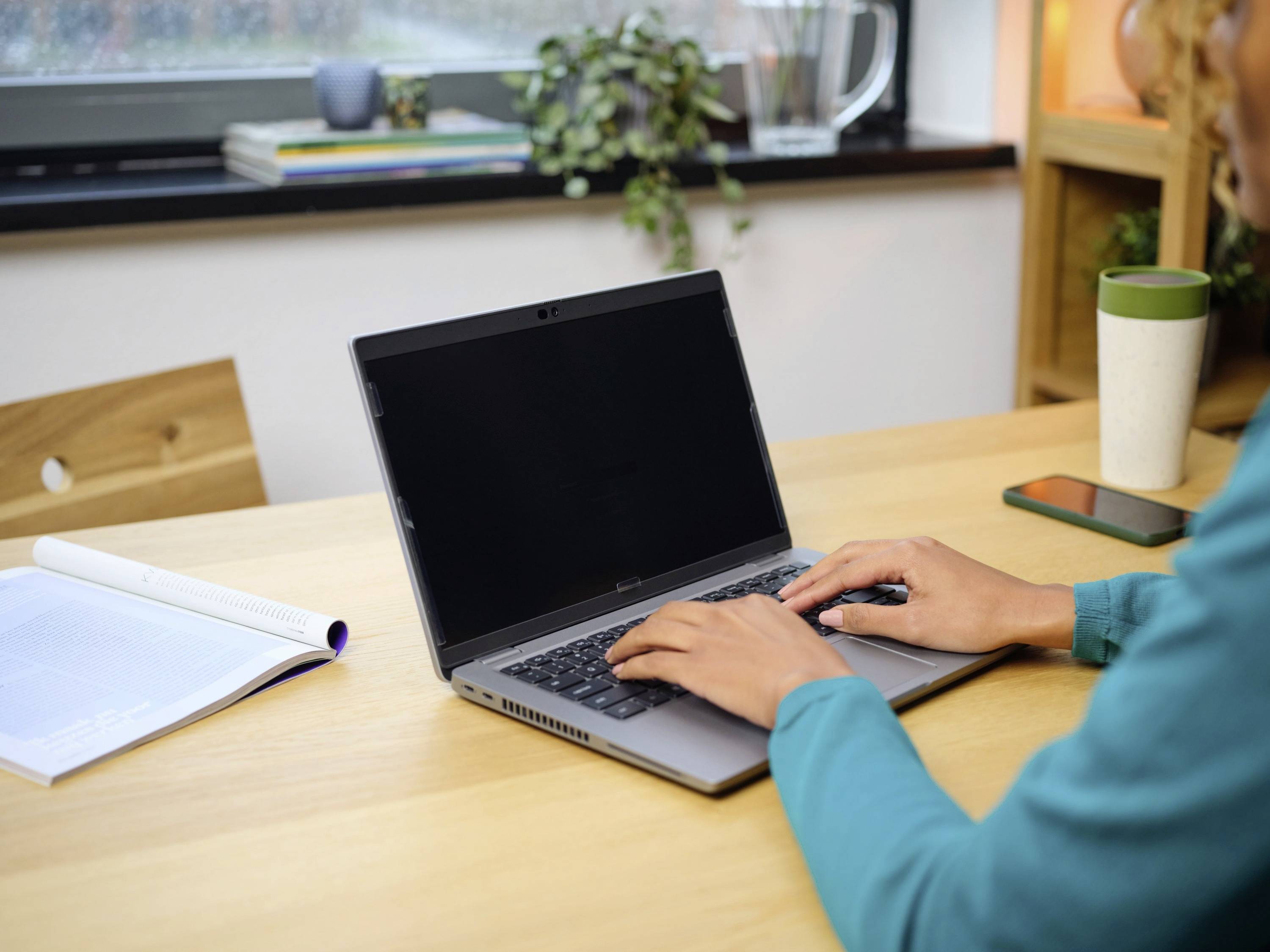 Persoon werkt op een laptop aan een houten tafel. Naast de laptop liggen een opengeslagen boek en een smartphone. Op de achtergrond zijn planten zichtbaar.