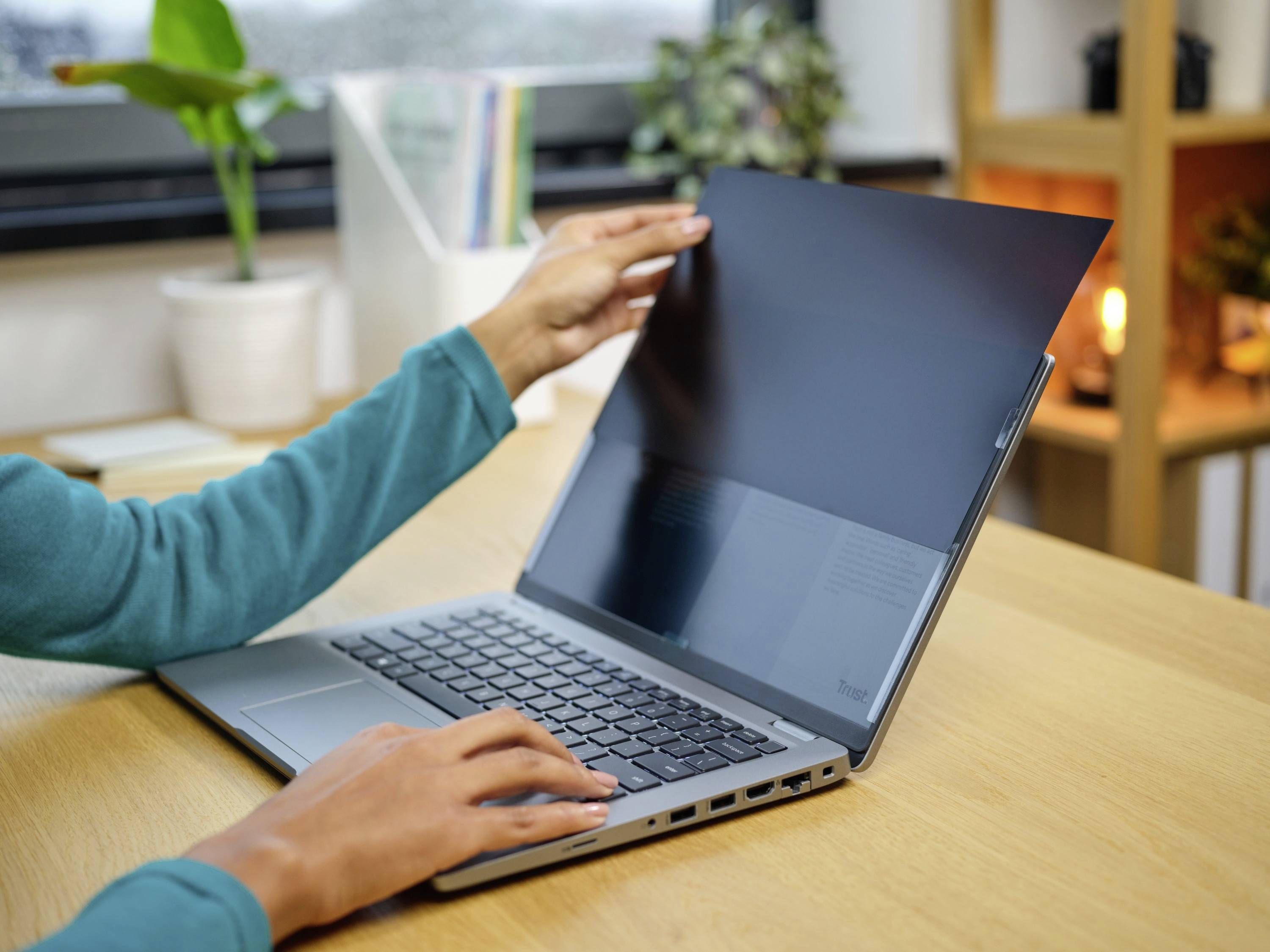 Een persoon opent het scherm van een laptop op een bureau. Op de achtergrond zijn planten en een rek zichtbaar.