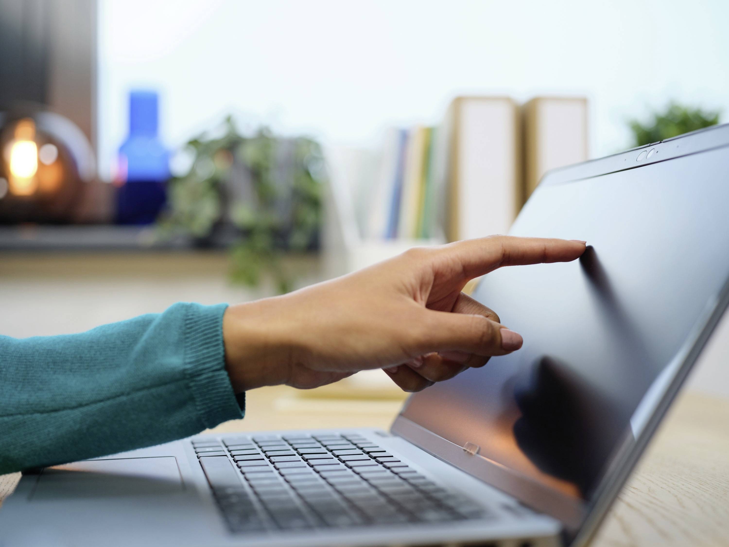 Hand wijst naar laptopscherm in een kantoor met planten op de achtergrond. Implicatie van technologie of digitale interactie.