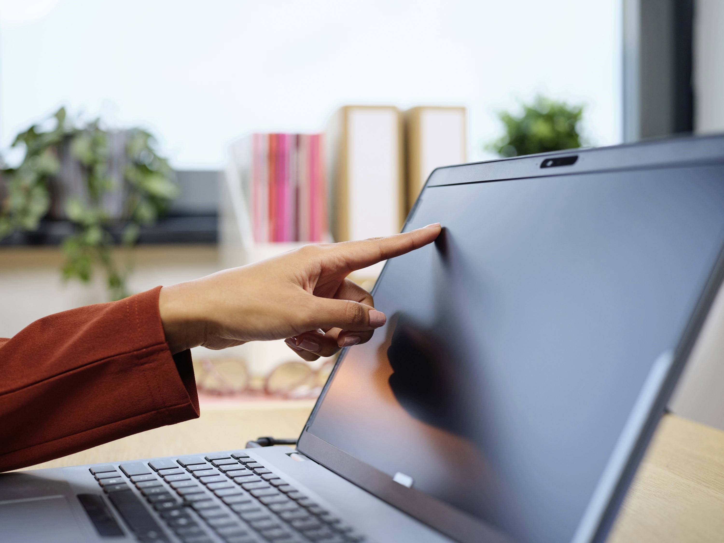 Een hand wijst naar het scherm van een laptop in een kantoor met planten en mappen op de achtergrond.