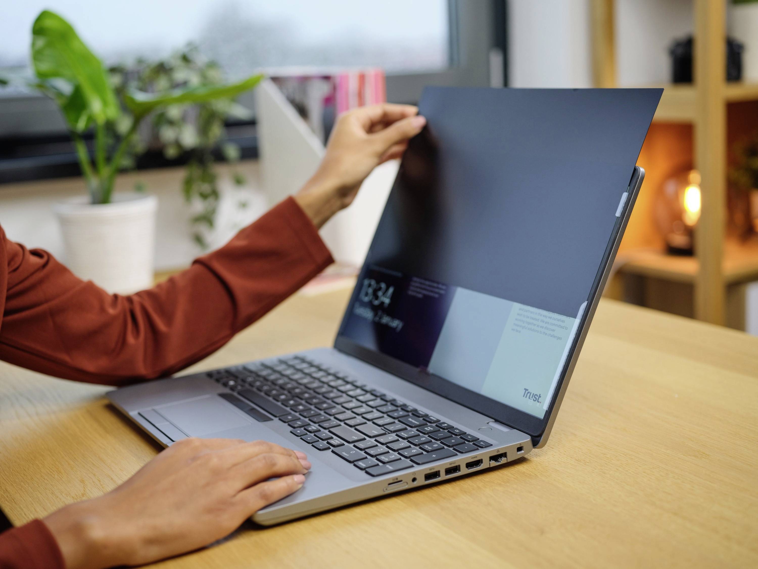 Een persoon opent een laptop aan een tafel. Op de achtergrond bevinden zich planten en boekenkasten die een gezellige kantooromgeving laten zien.