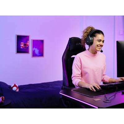 Een persoon met koptelefoon zit in een gamingstoel voor een computer, glimlachend. De ruimte is gehuld in paars licht.