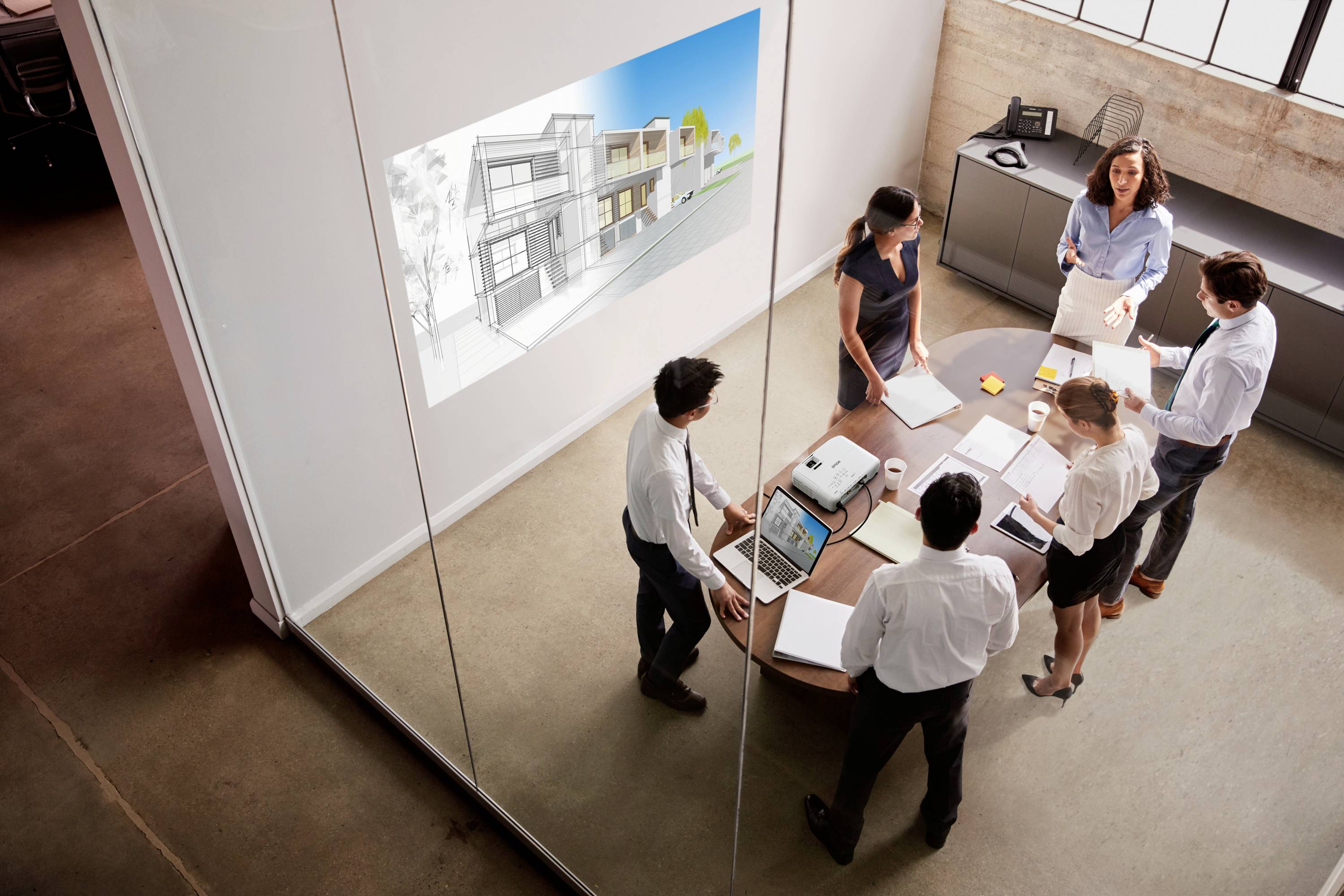 Een team van zes personen in formele kleding staat om een tafel in een modern kantoor. Een architectuurontwerp-projectie is zichtbaar aan de muur.