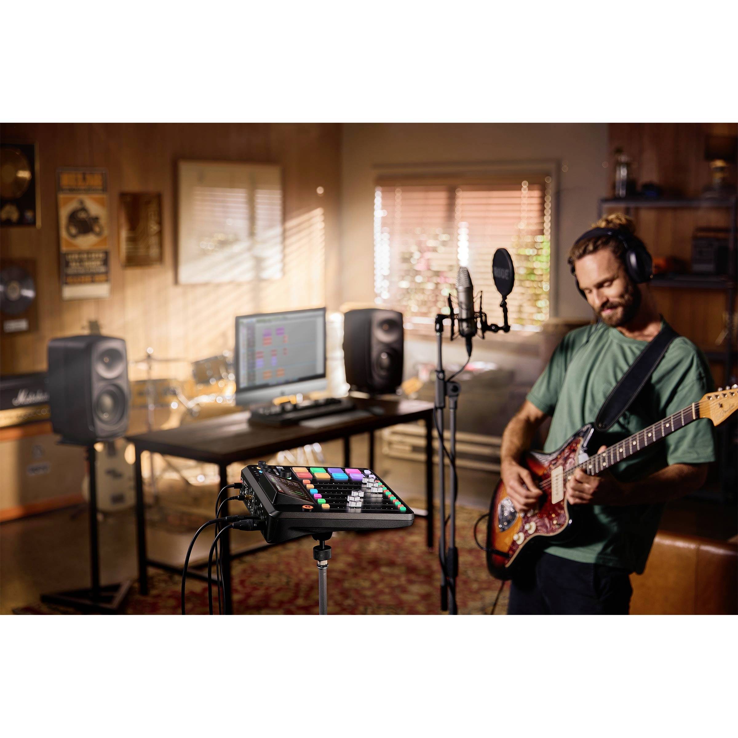 Een man speelt gitaar in een thuisstudio. Voor hem staat een microfoon, op de achtergrond een bureau met monitors en luidsprekers.