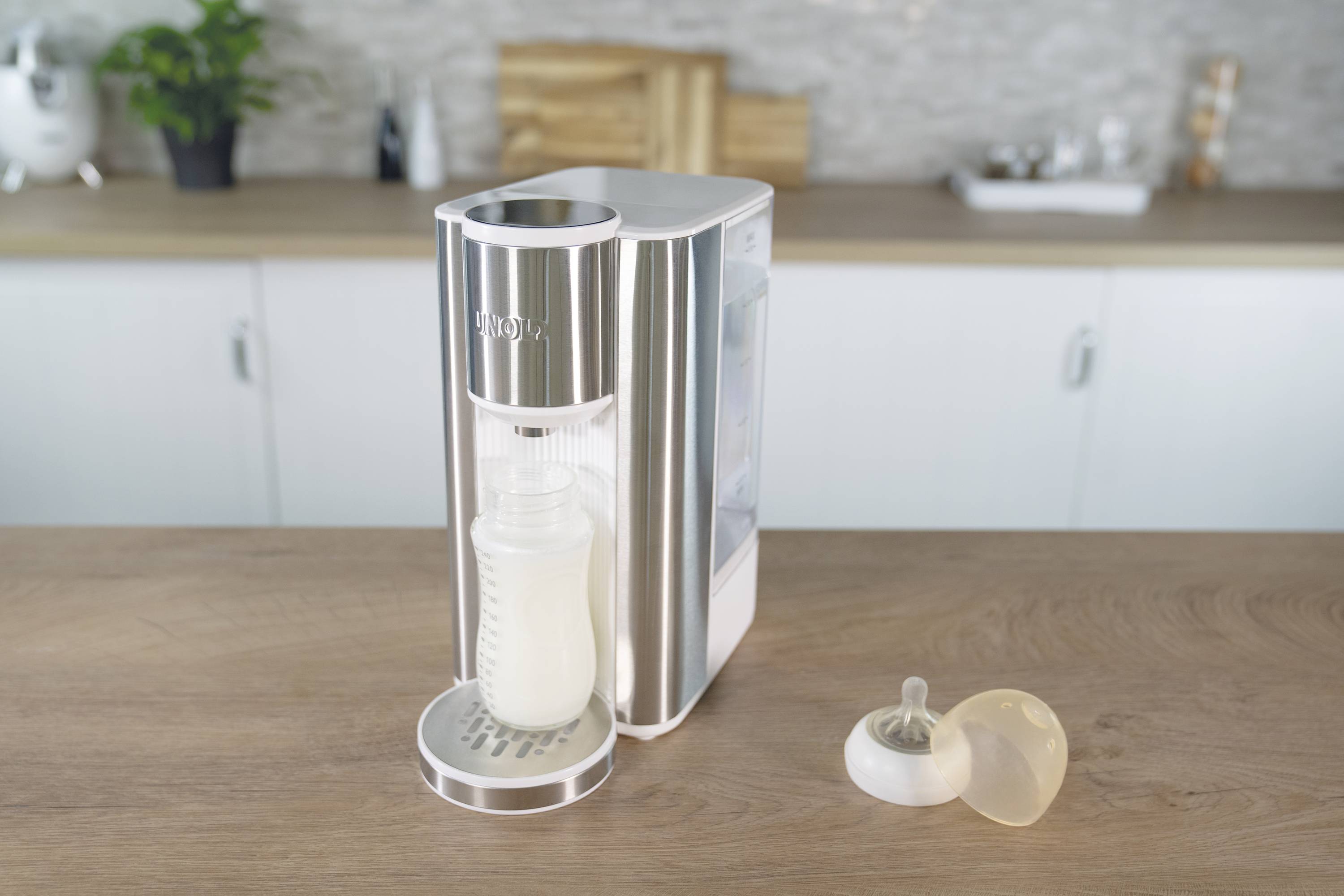 Een babyflesmachine op een aanrecht met naast zich een gevulde melkcontainer. Op de achtergrond bevindt zich een keukeninrichting.