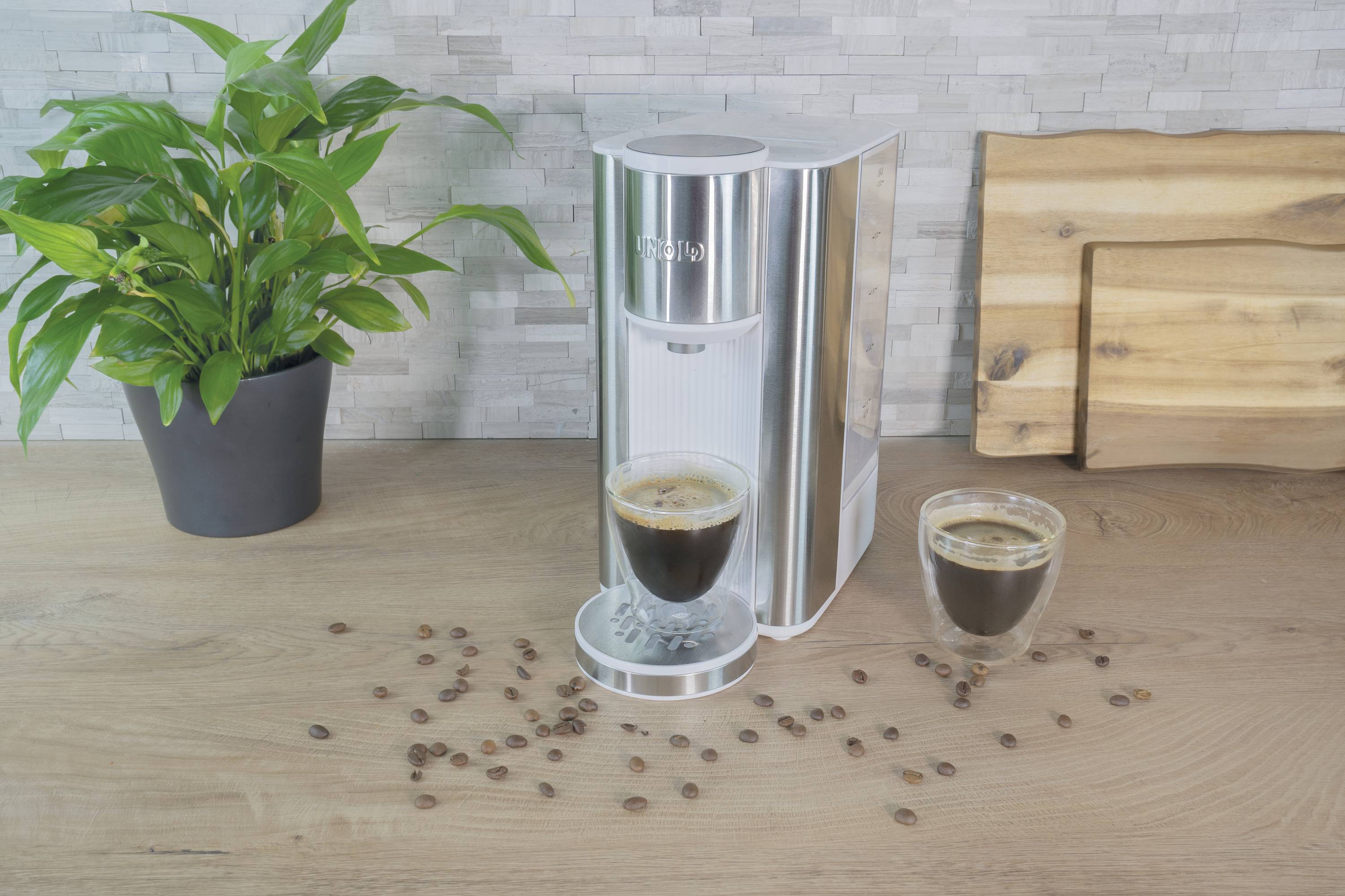 Een zilveren koffiemachine op een houten tafel naast een groene plant. Ernaast een kopje gevulde koffie en verspreid liggende bonen.