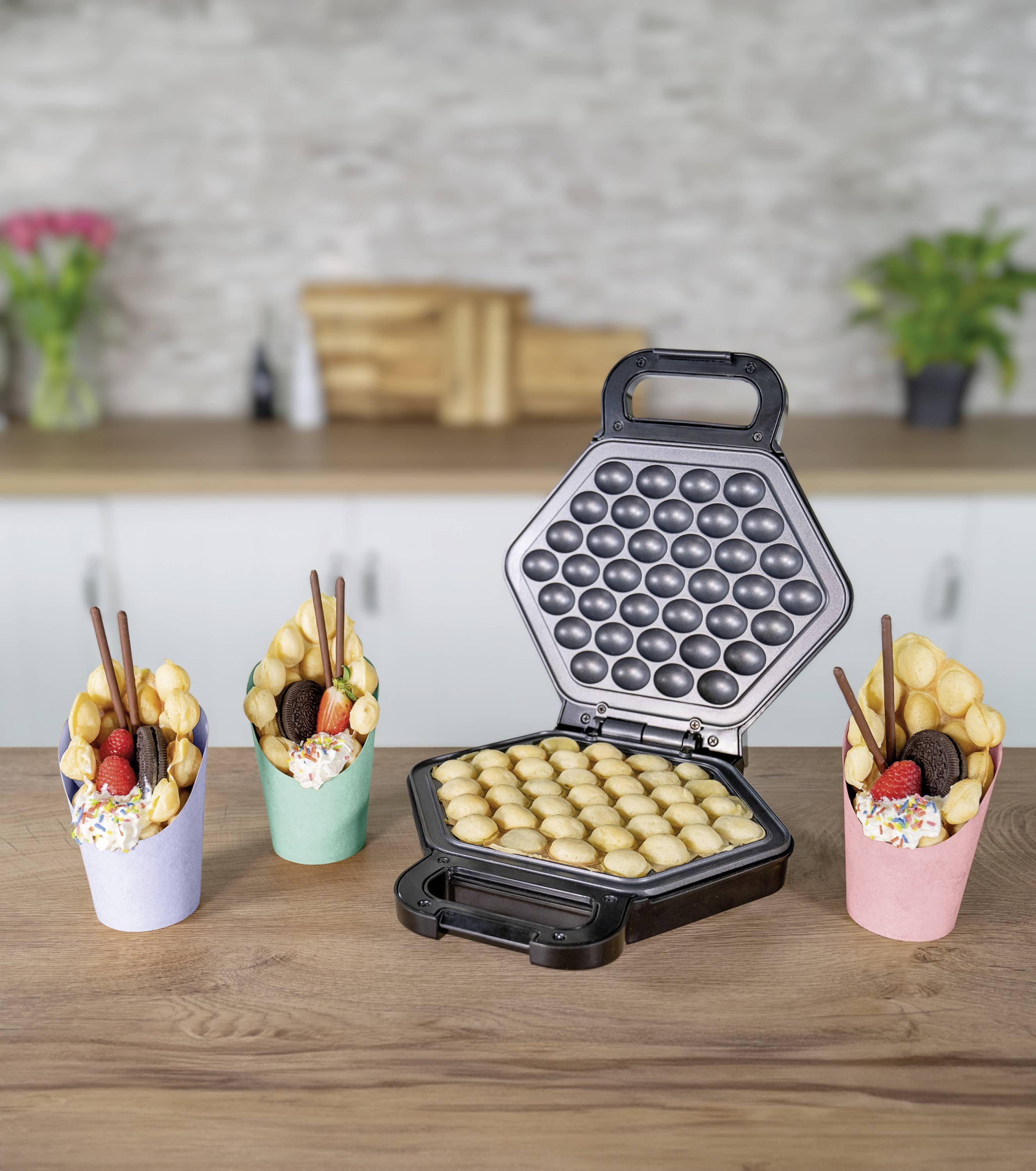 Een bubble wafel-ijzer met goudbruine wafels. Ernaast drie afgewerkte bubble wafels in kleurrijke bekers, gevuld met fruit en snoep.