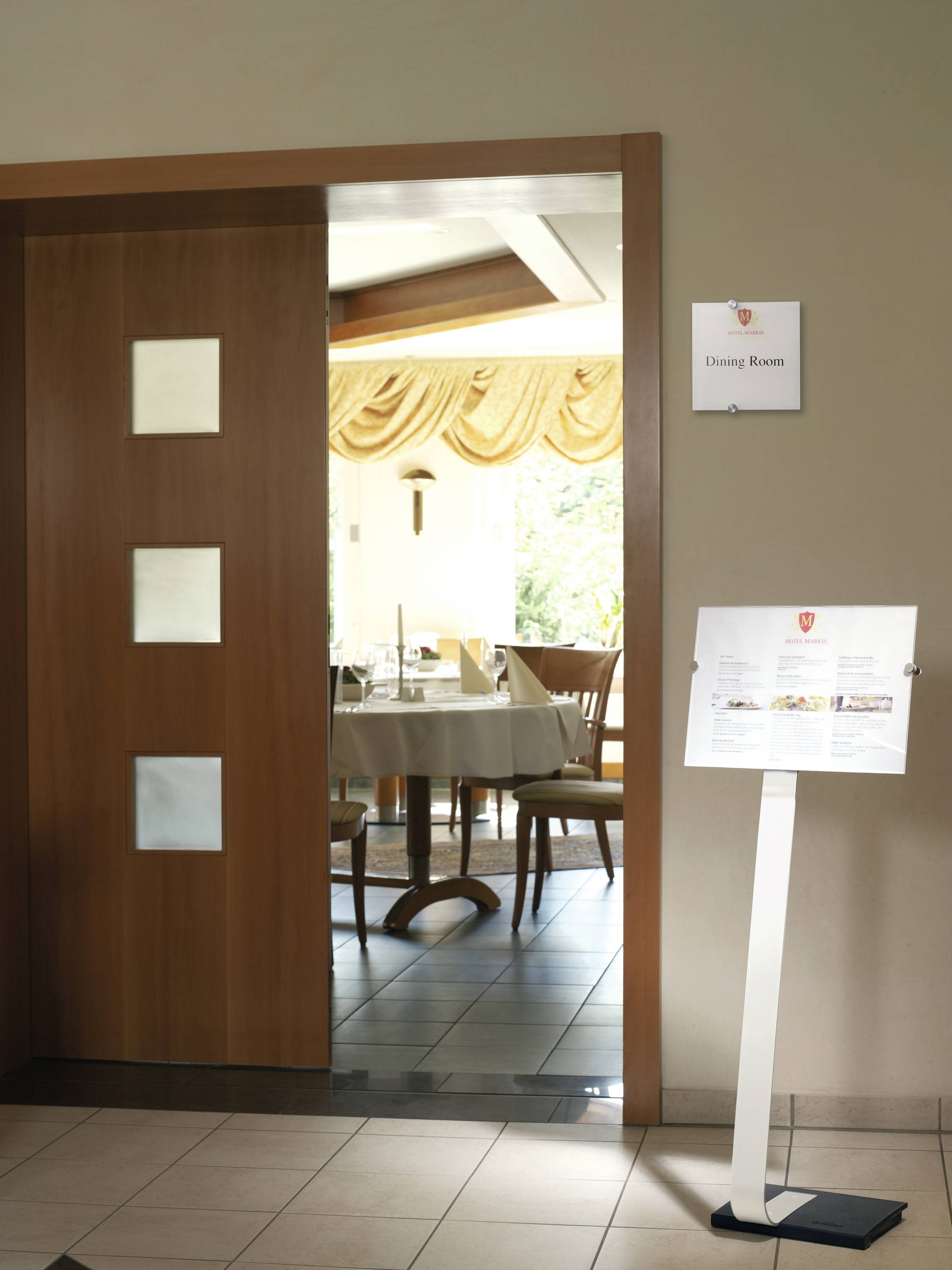 Ingang naar een eetzaal met een houten schuifdeur en een lessenaar. Zicht op gedekte tafels aan de binnenkant. Bord: 'Eetzaal'.