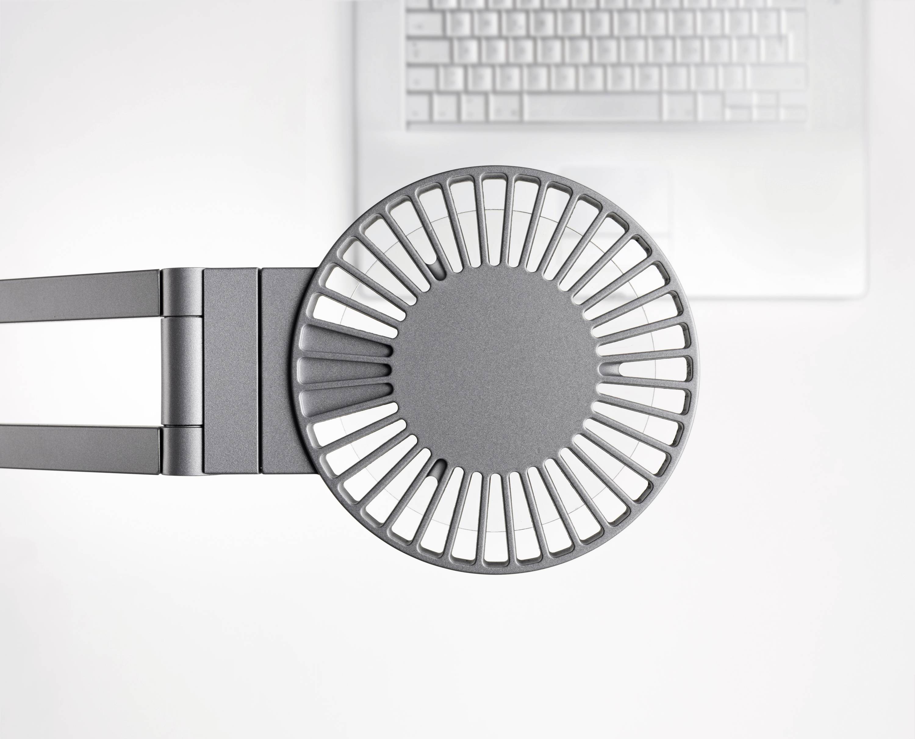 Een ronde, grijze bureauventilator met rooster, van bovenaf gezien, staat voor een wit computertoetsenbord op een bureau.