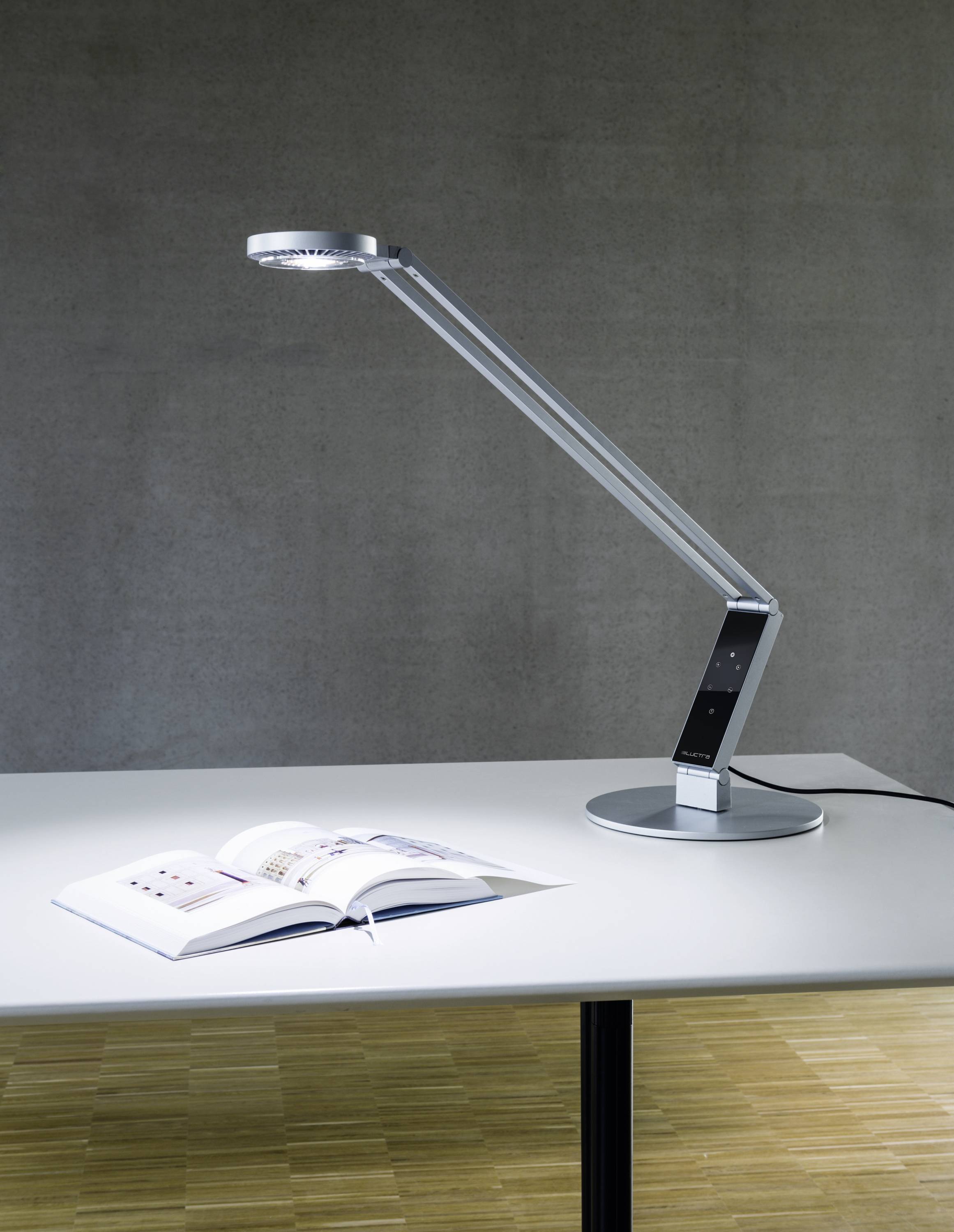 Een moderne bureaulamp met een lange, slanke arm verlicht een opengeslagen boek op een witte tafel voor een grijze muur.