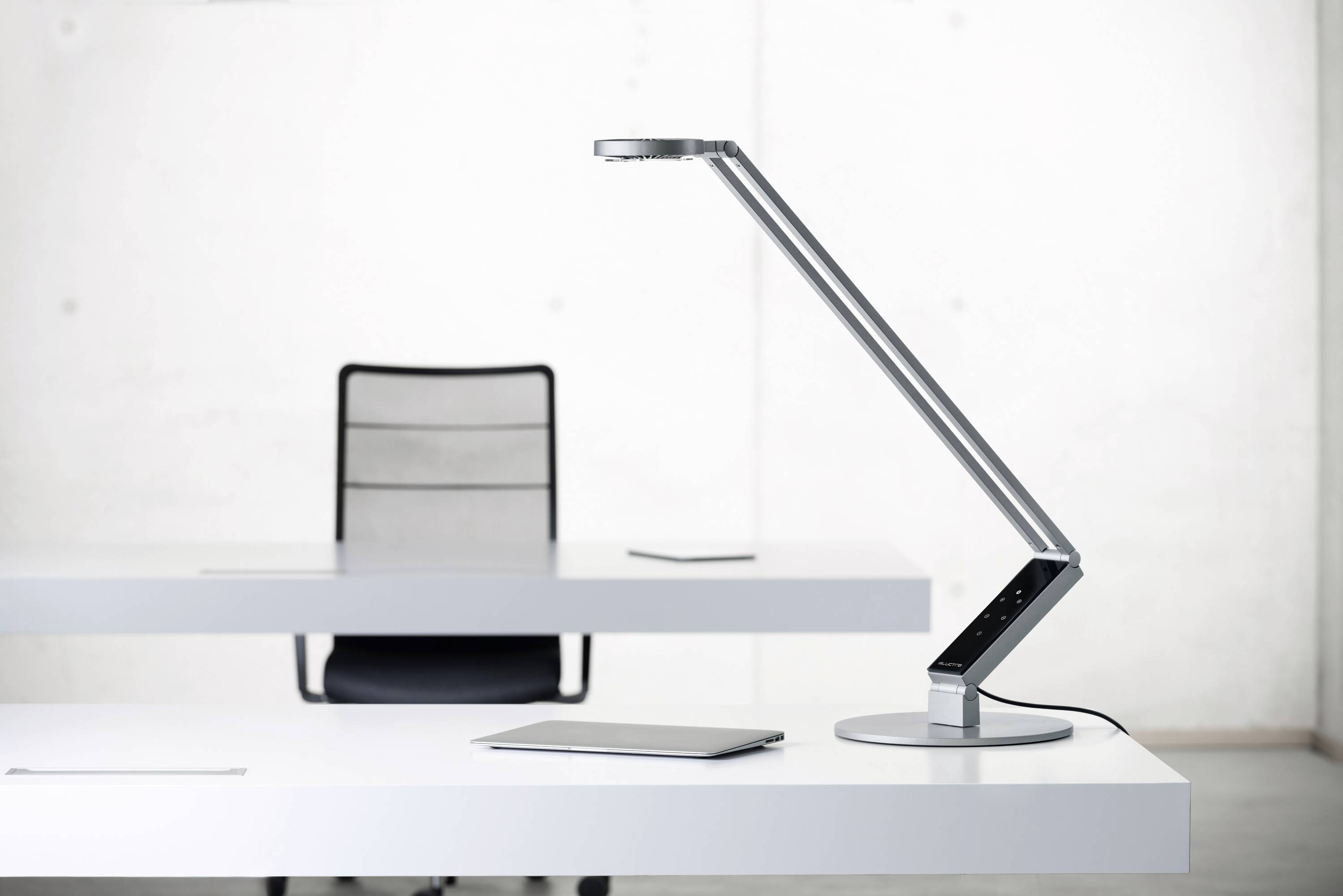 Een moderne, verstelbare bureaulamp staat op een wit bureau, op de achtergrond een zwarte bureaustoel.