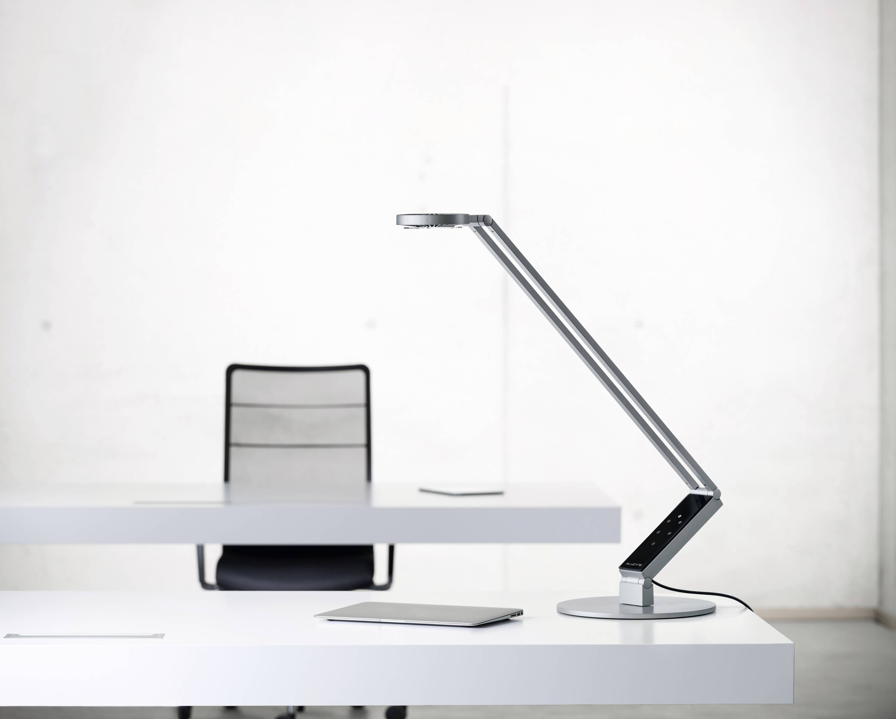 Moderne bureaulamp op een witte tafel, naast een tablet. Op de achtergrond staat een lege stoel.