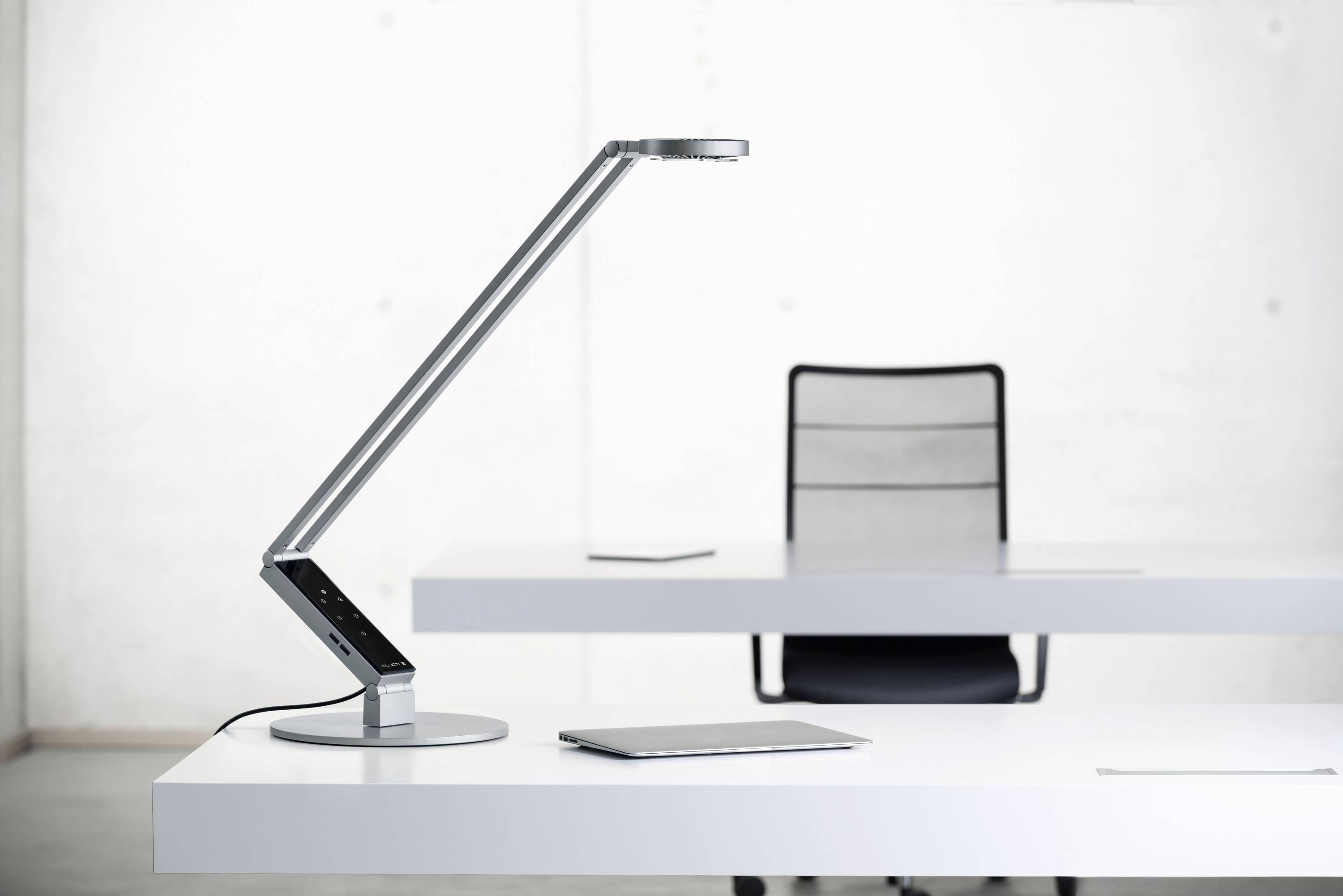 Moderne bureaulamp naast laptop en tablet op een witte tafel. Achtergrond toont lege bureaustoel en lichte muur.