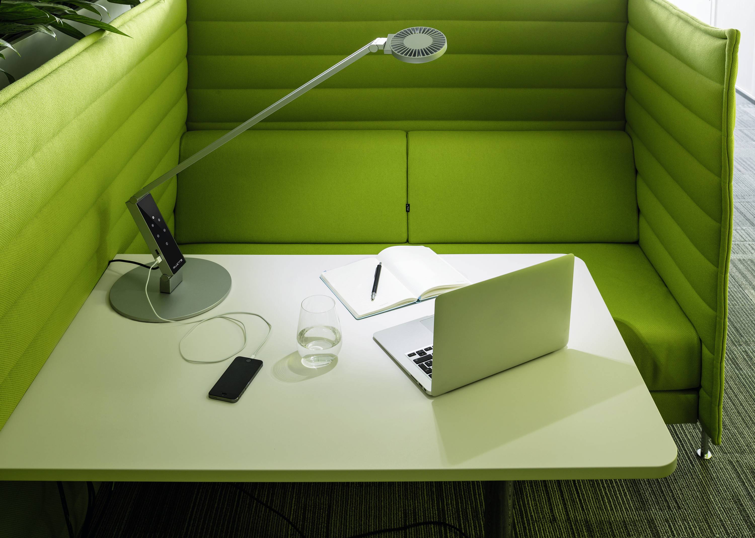 Een moderne bureautafel met een laptop, een opengeslagen notitieboek, een bureaulamp, een smartphone en een glas water op een groene bank of zitgelegenheid.