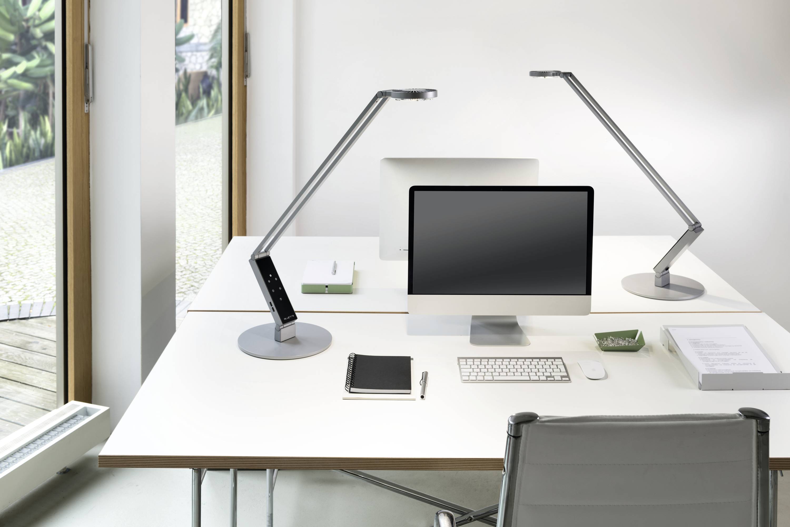 Kantoor met minimalistisch design: Twee bureaus met iMac, toetsenbord, muis, documenten en moderne bureaulampen. Raam op de achtergrond.