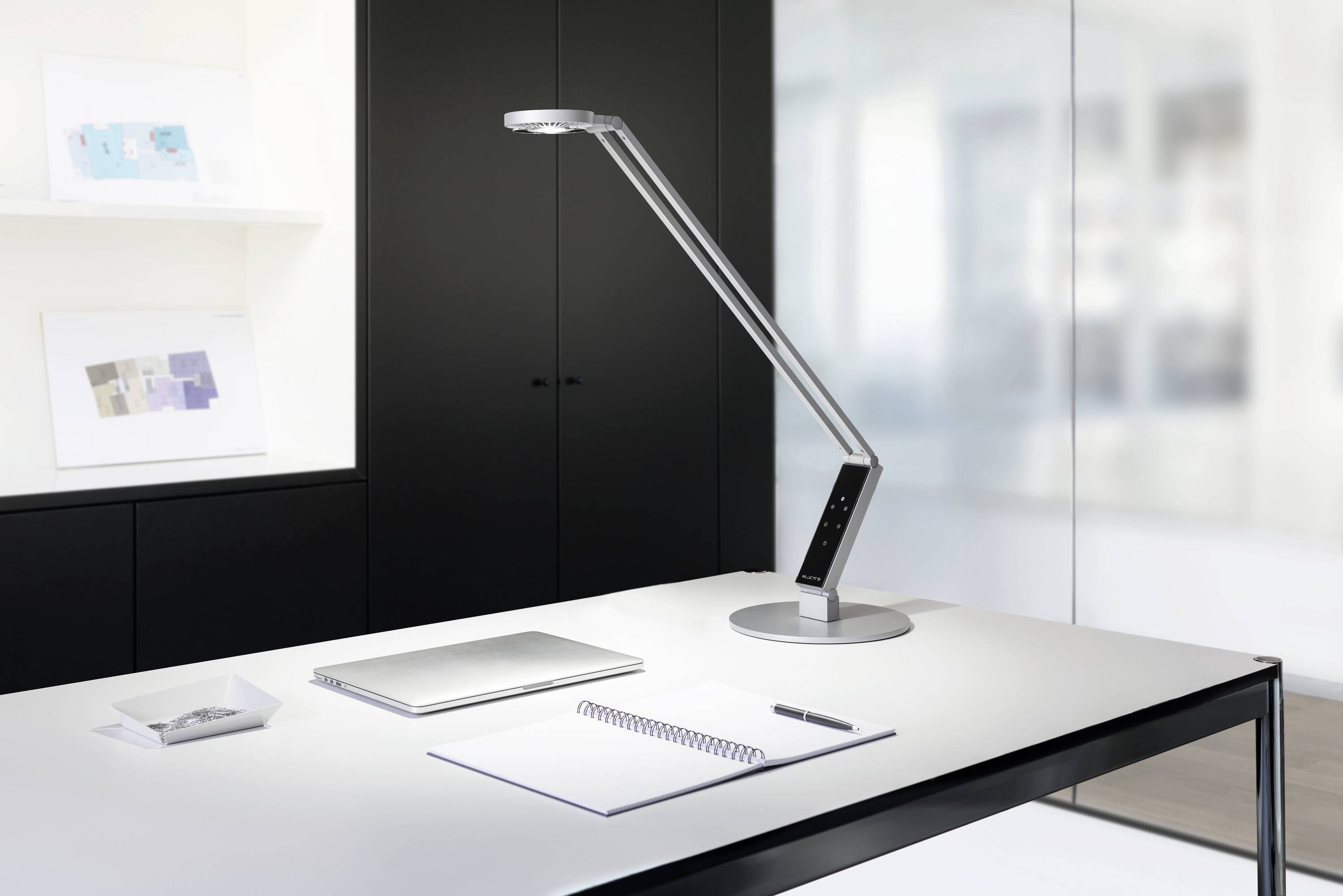 Een moderne bureau met een laptop, notitieboekje en pen. Een slanke bureaulamp verlicht de werkplek in de lichte ruimte.