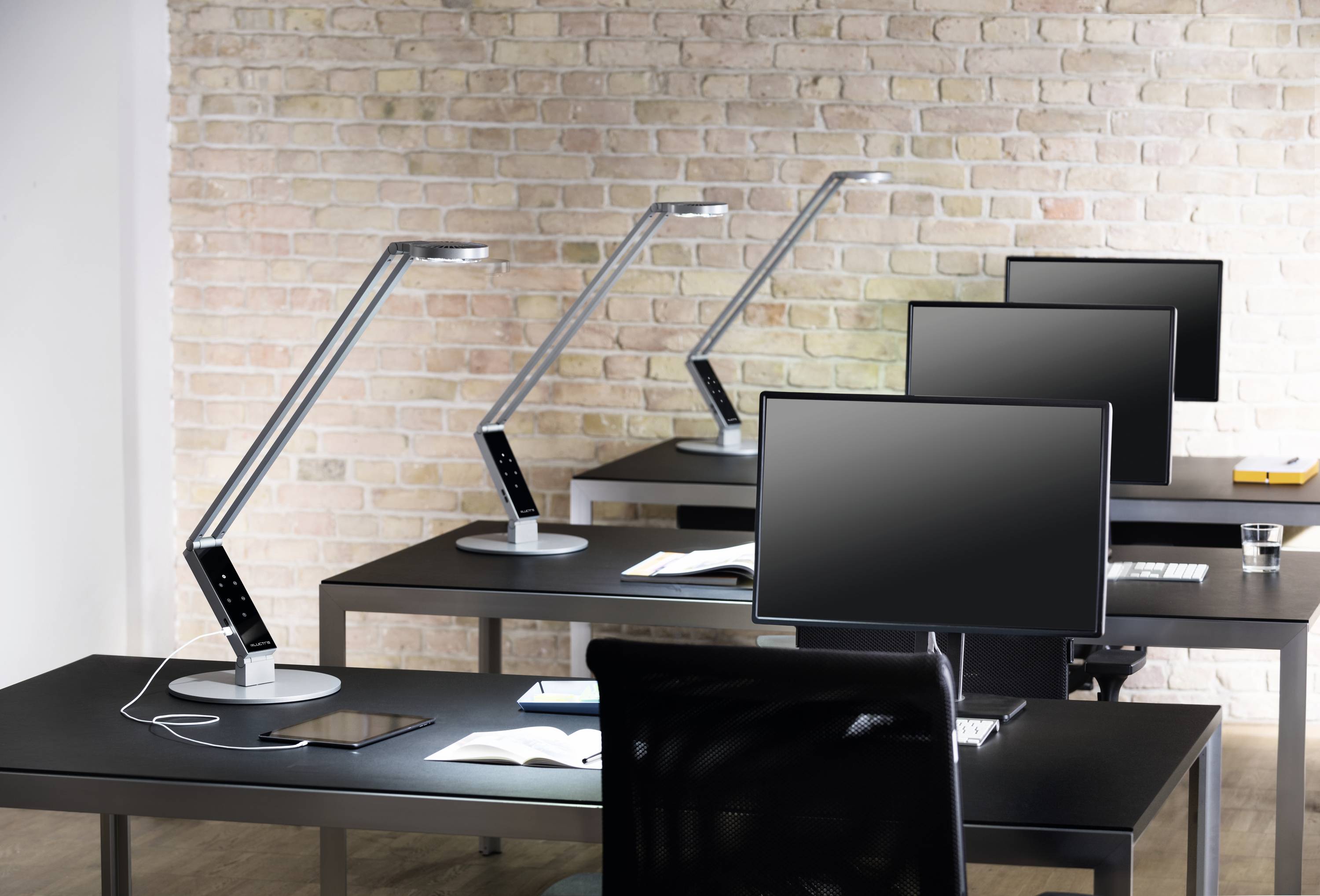 Verschillende lege werkplekken in een modern kantoor met zwarte bureaus, monitors en verstelbare lampen voor een bakstenen muur.