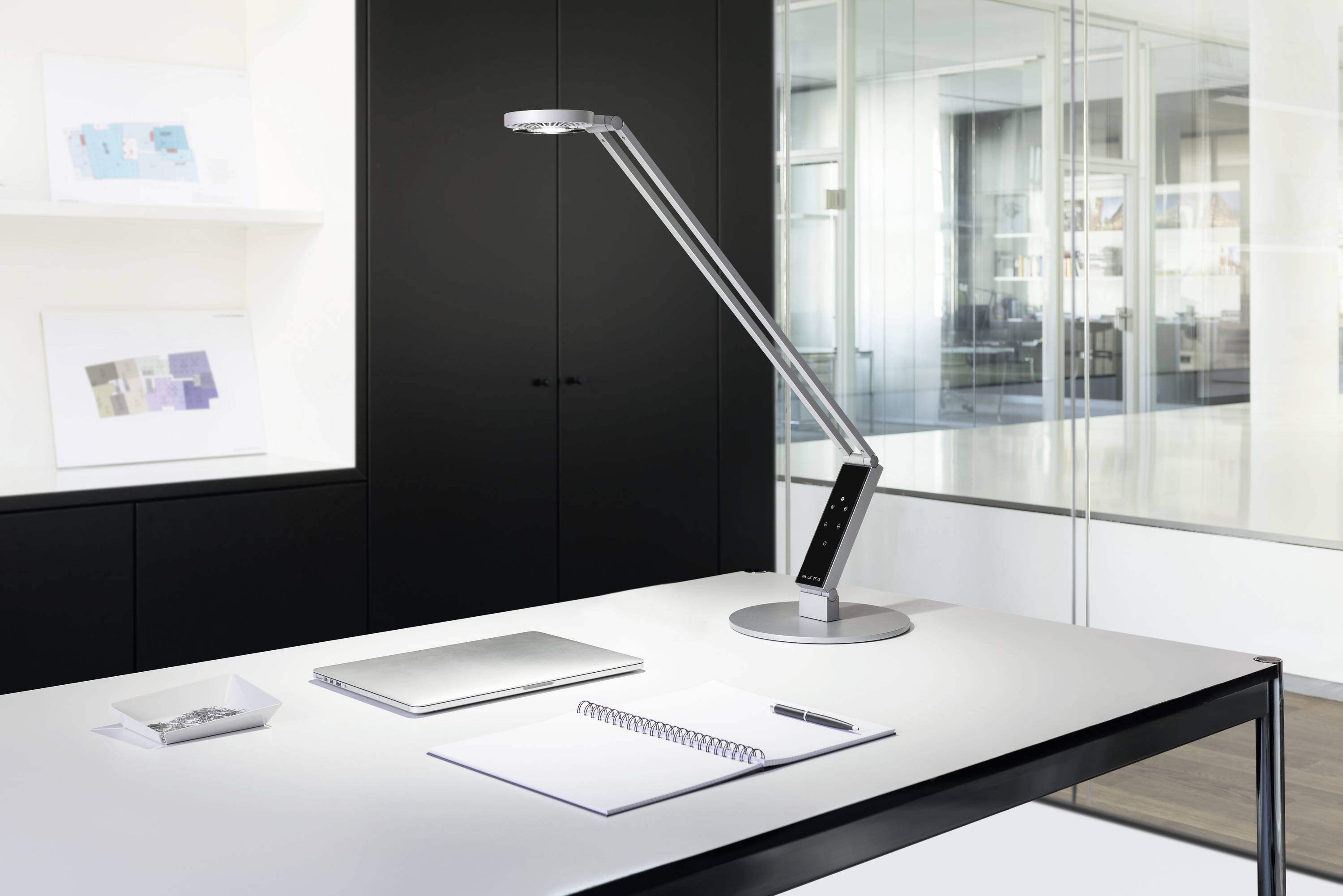 Een modern kantoor met een tafel waarop een laptop, notitieboek en pen liggen. Een slanke bureaulamp verlicht de werkplek.