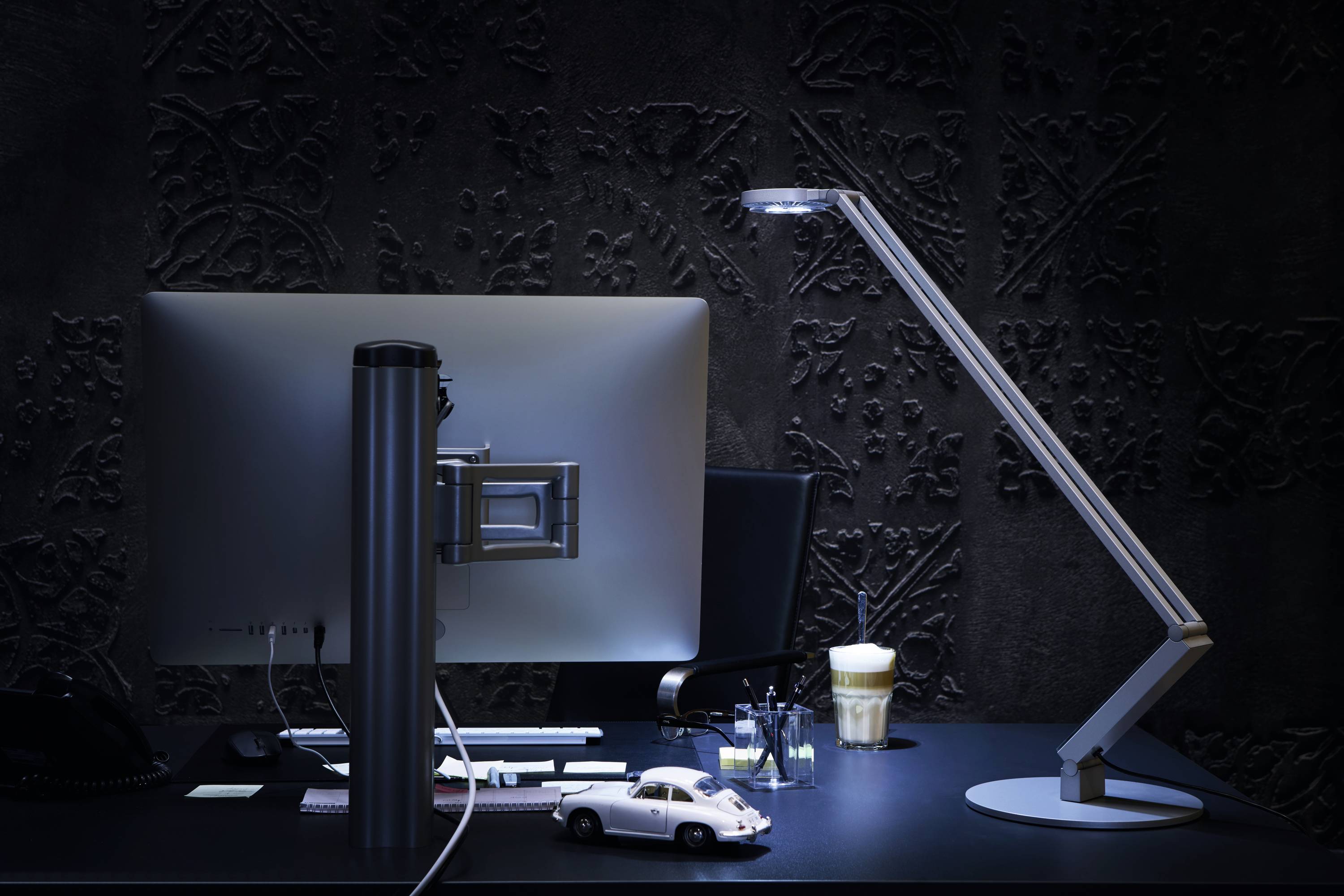 Een bureau met een grote monitor, een verstelbare lamp, een speelgoedauto, pennen en een drankje. Donkere, elegante achtergrond.