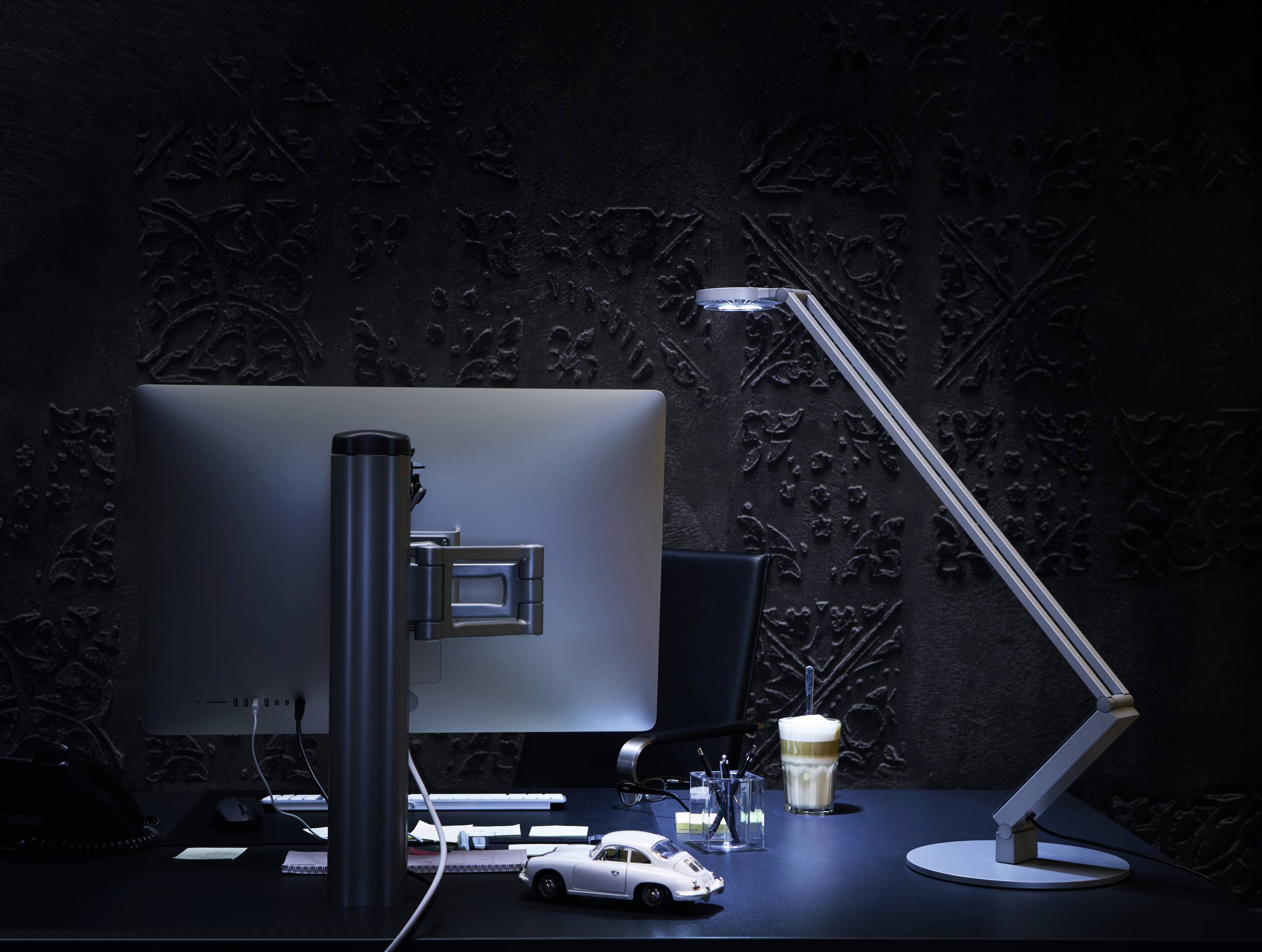 Een moderne bureau met een beeldscherm, een lamp, een modelauto en een drankje op een donkere, decoratieve achtergrond.