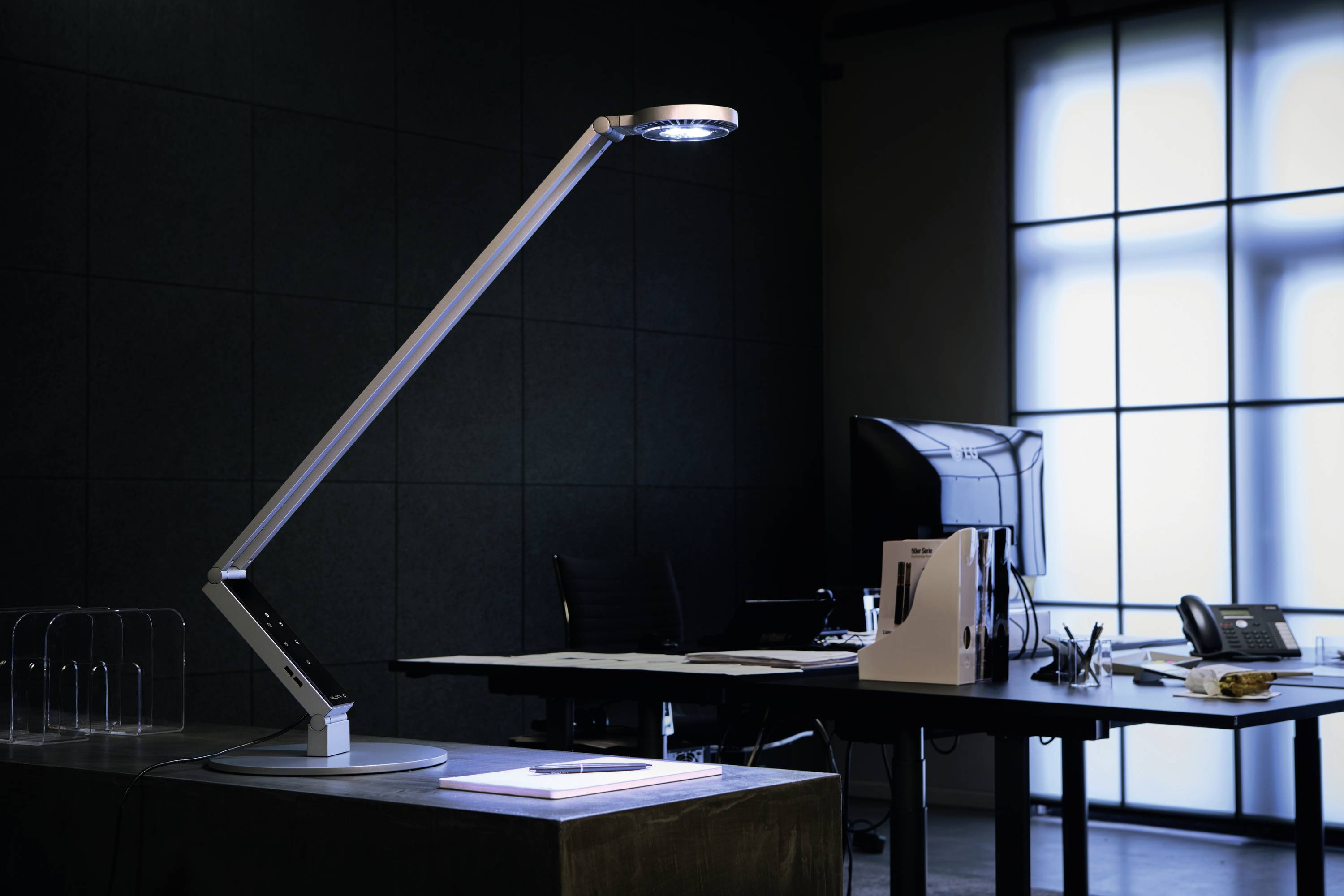 Een bureaulamp verlicht een tafel met een laptop in een modern kantoor. Op de achtergrond zijn ramen en een monitor te zien.