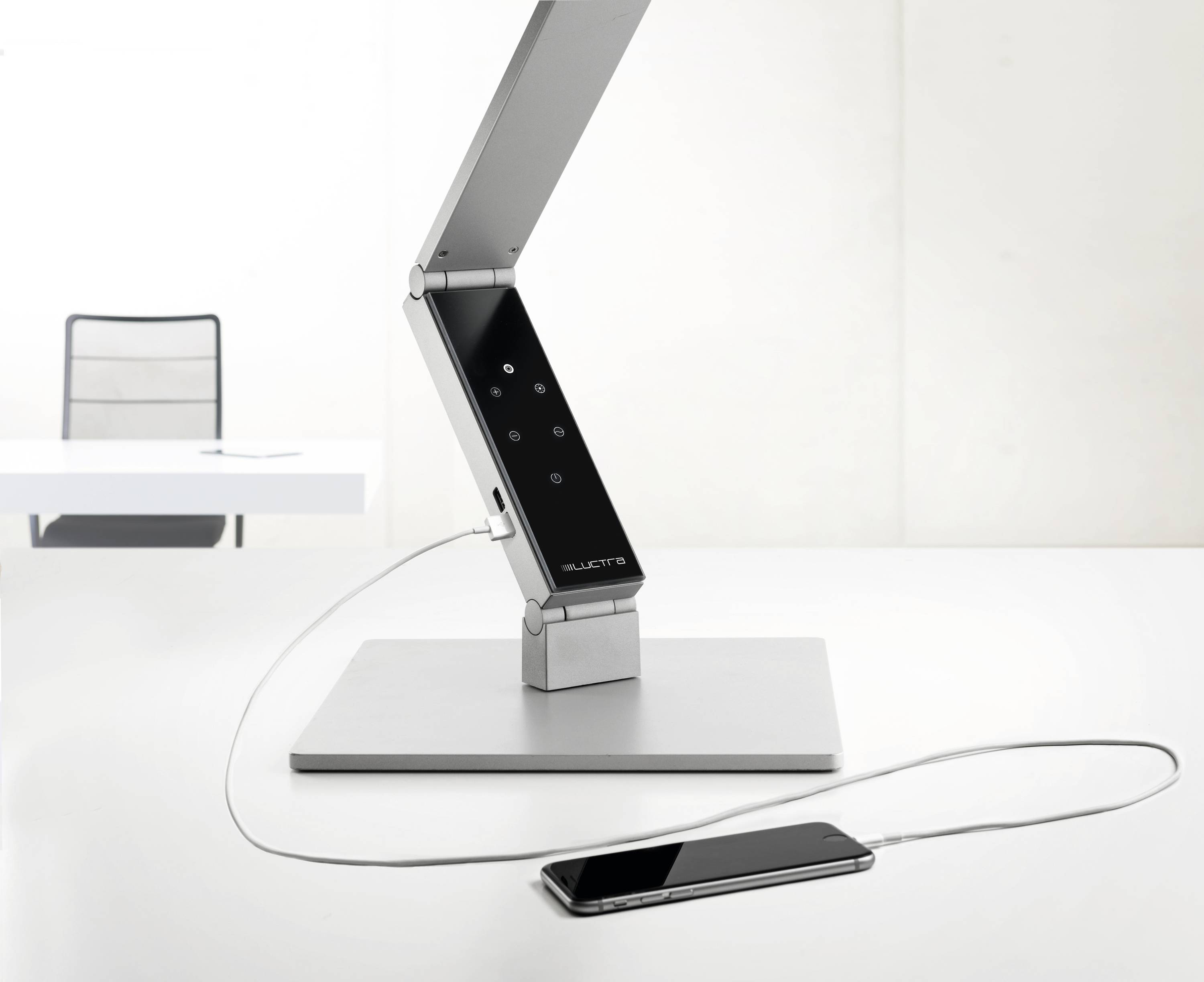 Een moderne bureaulamp met een verstelbare arm, geïntegreerd aanraakpaneel, verbonden met een smartphone via een oplaadkabel op een bureau.