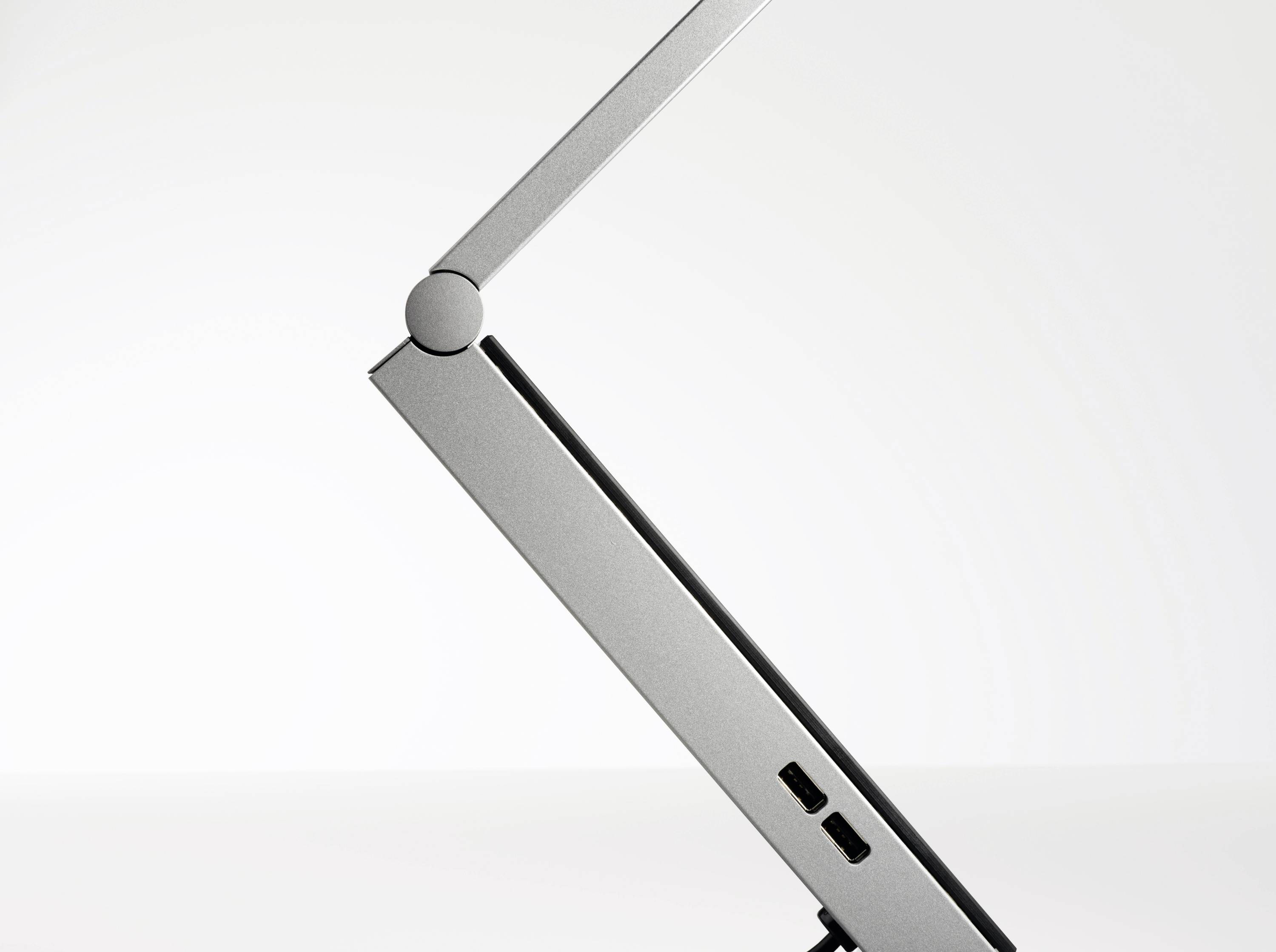 Een zijaanzicht van een zilveren, minimalistische bureaulamp met twee USB-poorten aan de voet, tegen een lichte achtergrond.
