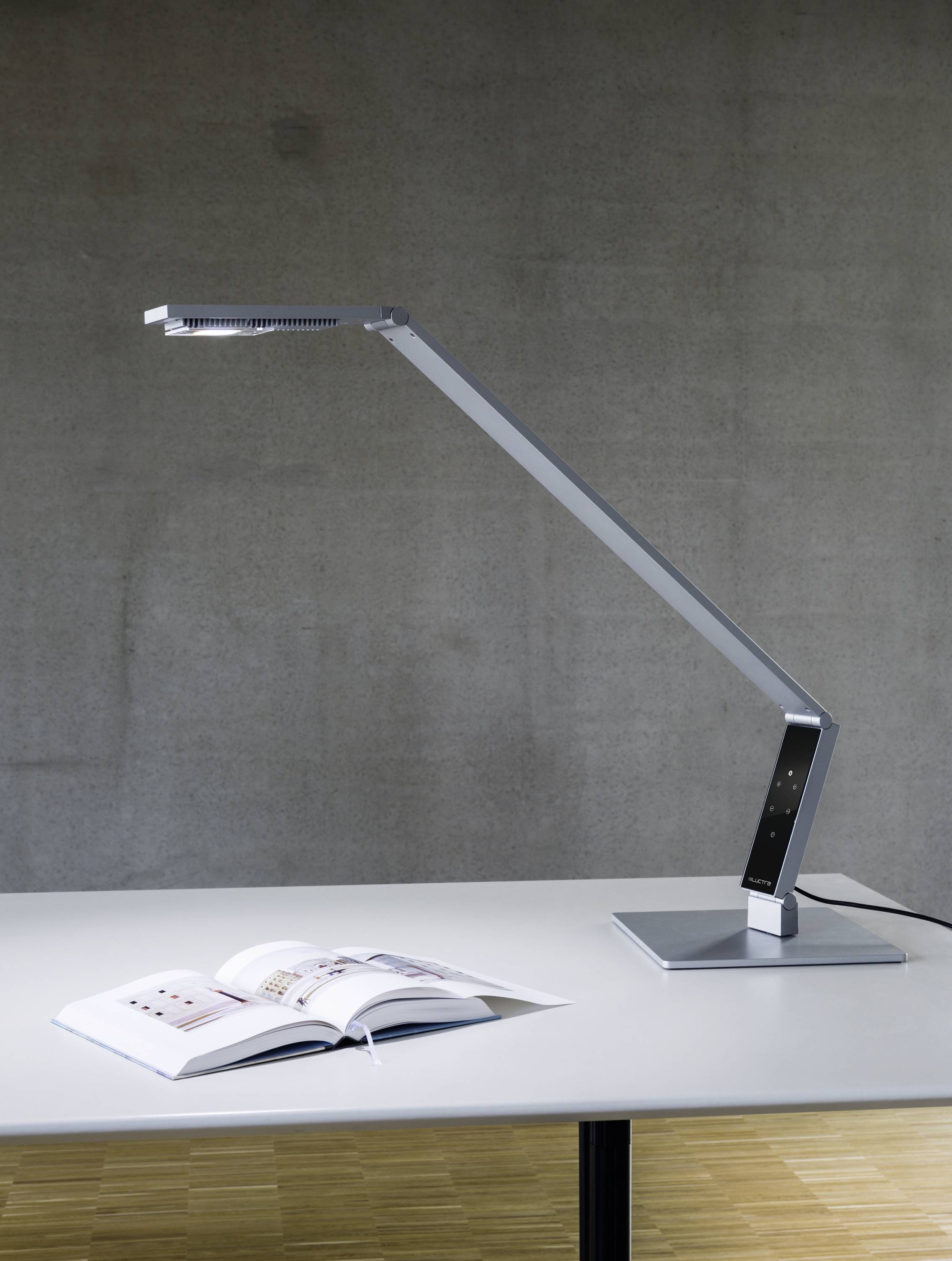 Een moderne, grijze bureaulamp verlicht een opengeslagen boek op een tafel. De achtergrond is een subtiele grijze muur.