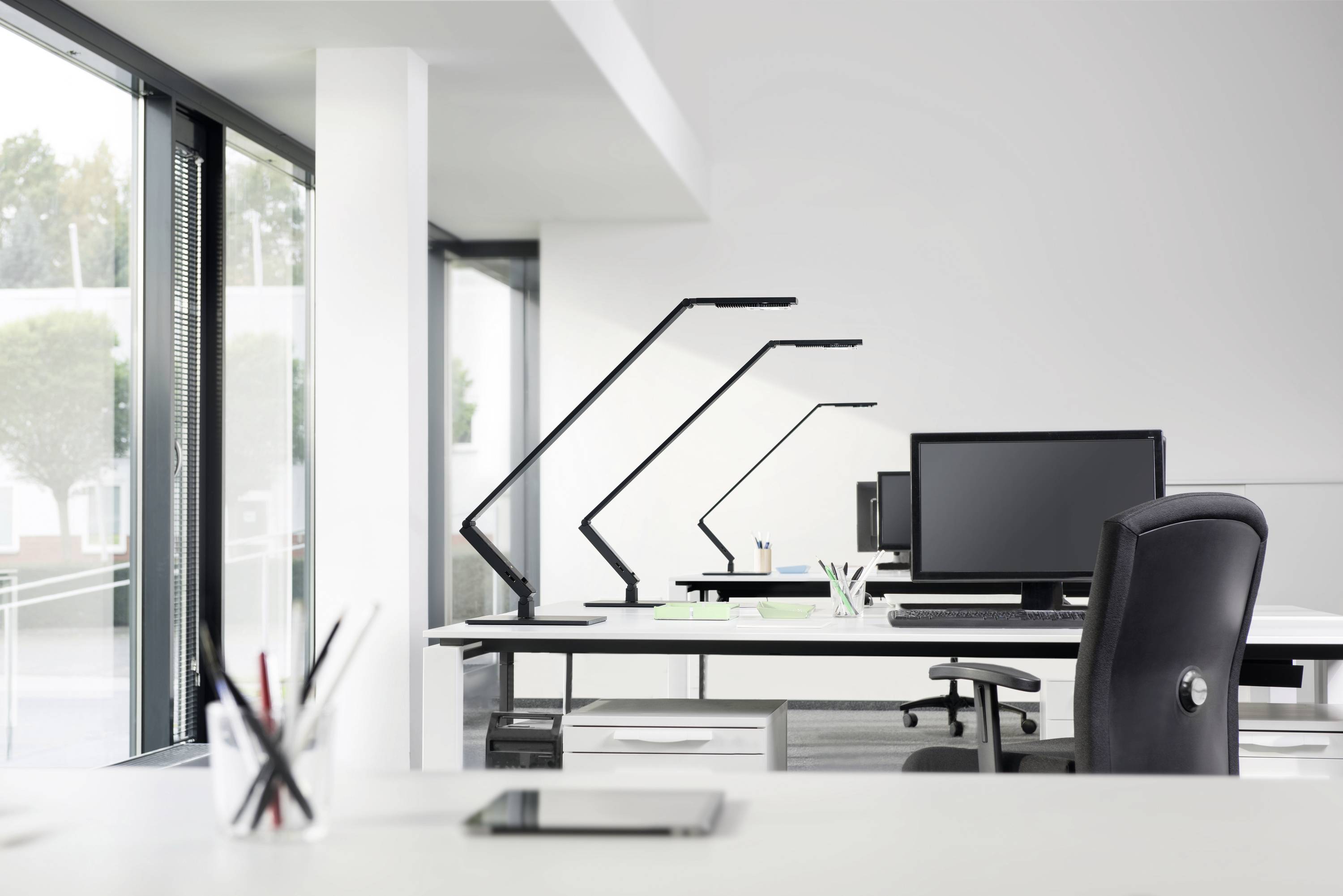 Een modern, goed verlicht kantoor met open ramen. Drie bureaus met monitors en stoelen zijn zichtbaar.