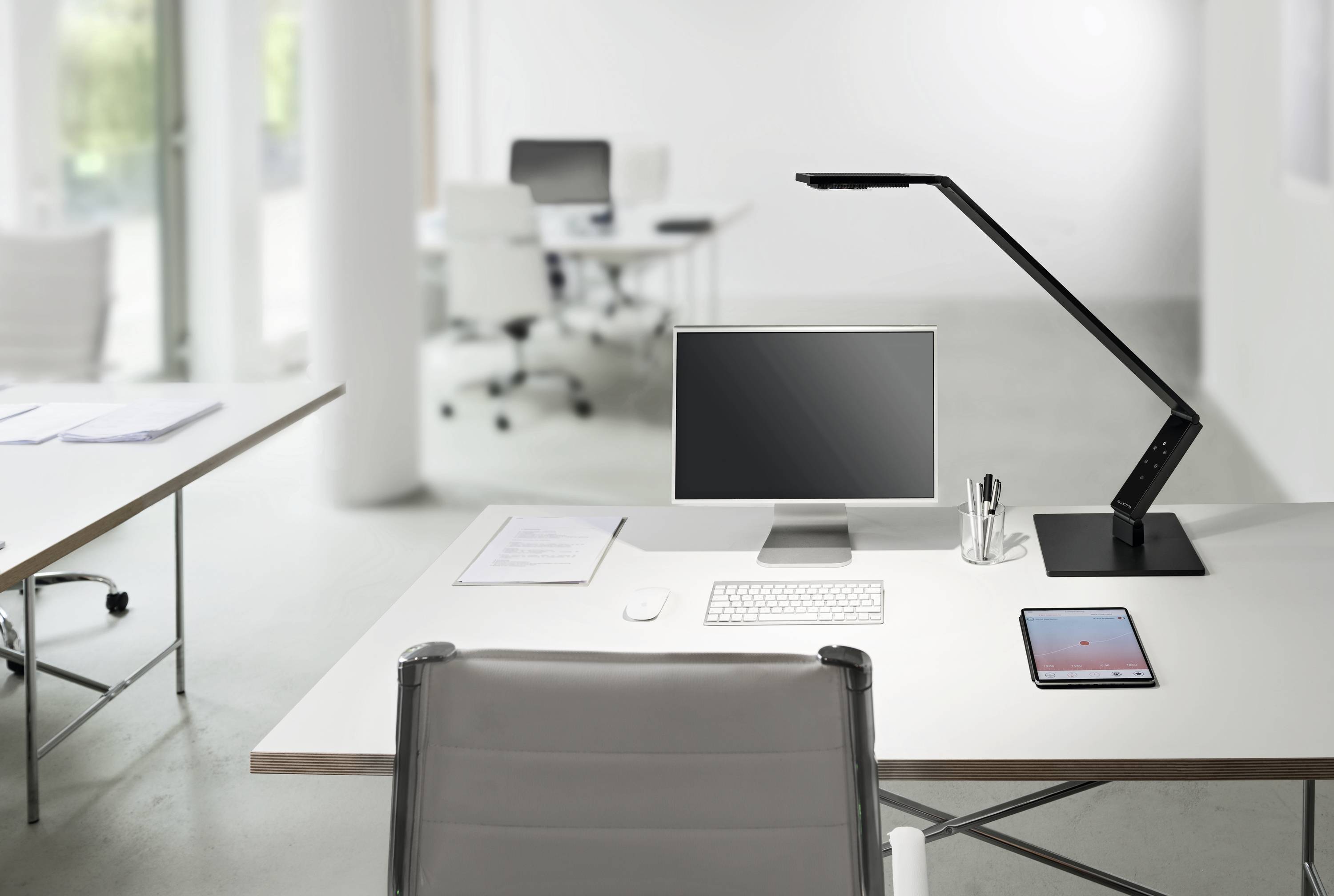 Een modern, licht kantoor met een bureau op de voorgrond. Op het bureau staan een monitor, een lamp en een tablet. Op de achtergrond zijn meerdere werkplekken zichtbaar.