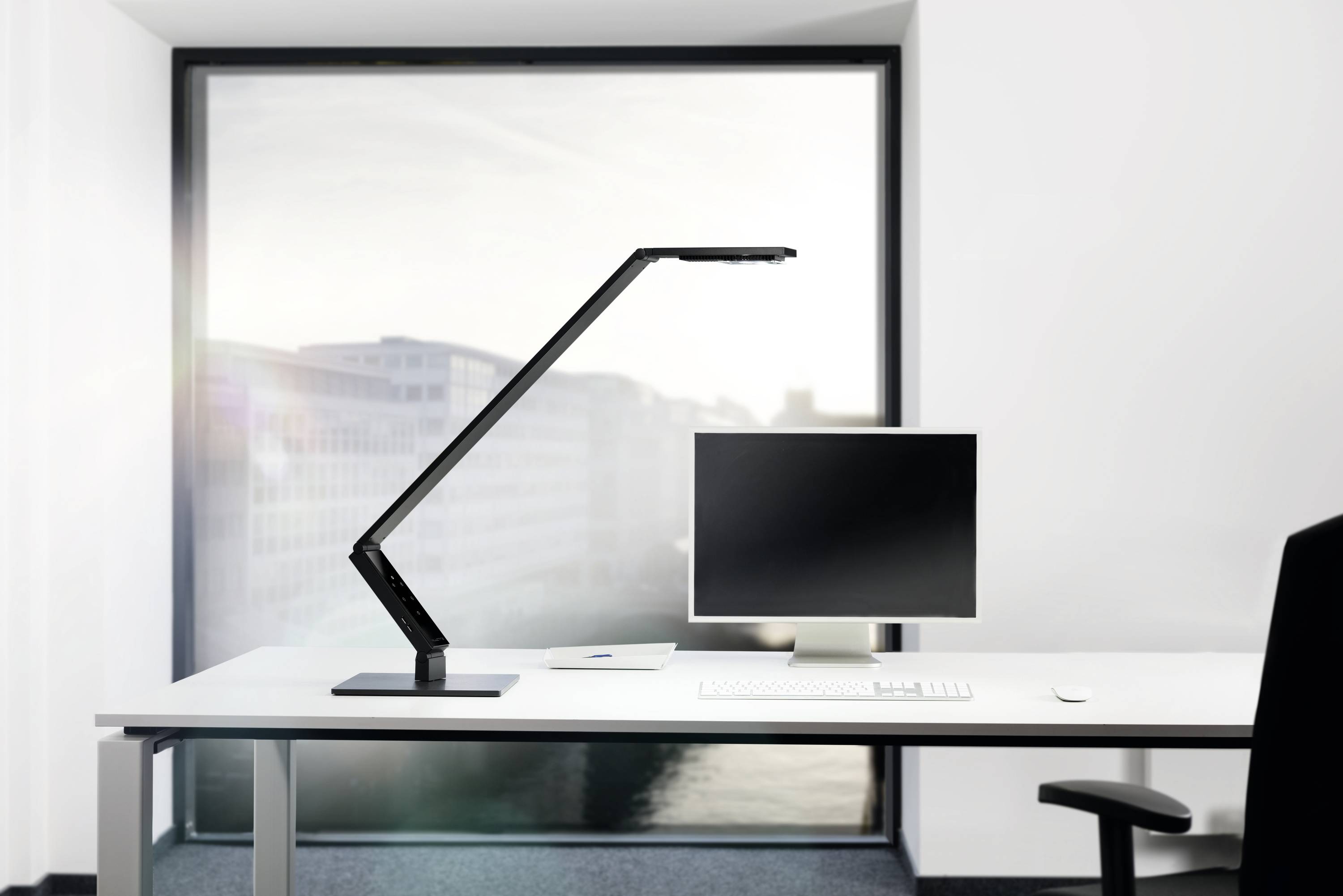 Een moderne werkplek met een bureau, een computerscherm, een slanke zwarte lamp en een groot raam op de achtergrond.