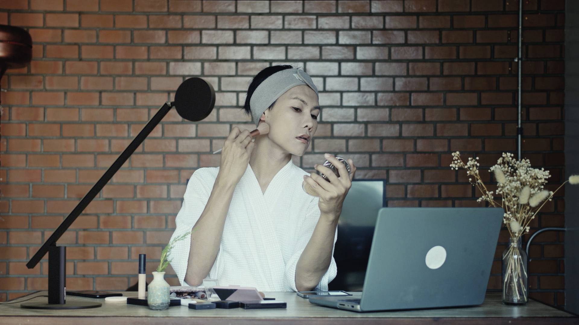 Een persoon in een badjas doet make-up voor een laptop, met een microfoon op tafel.