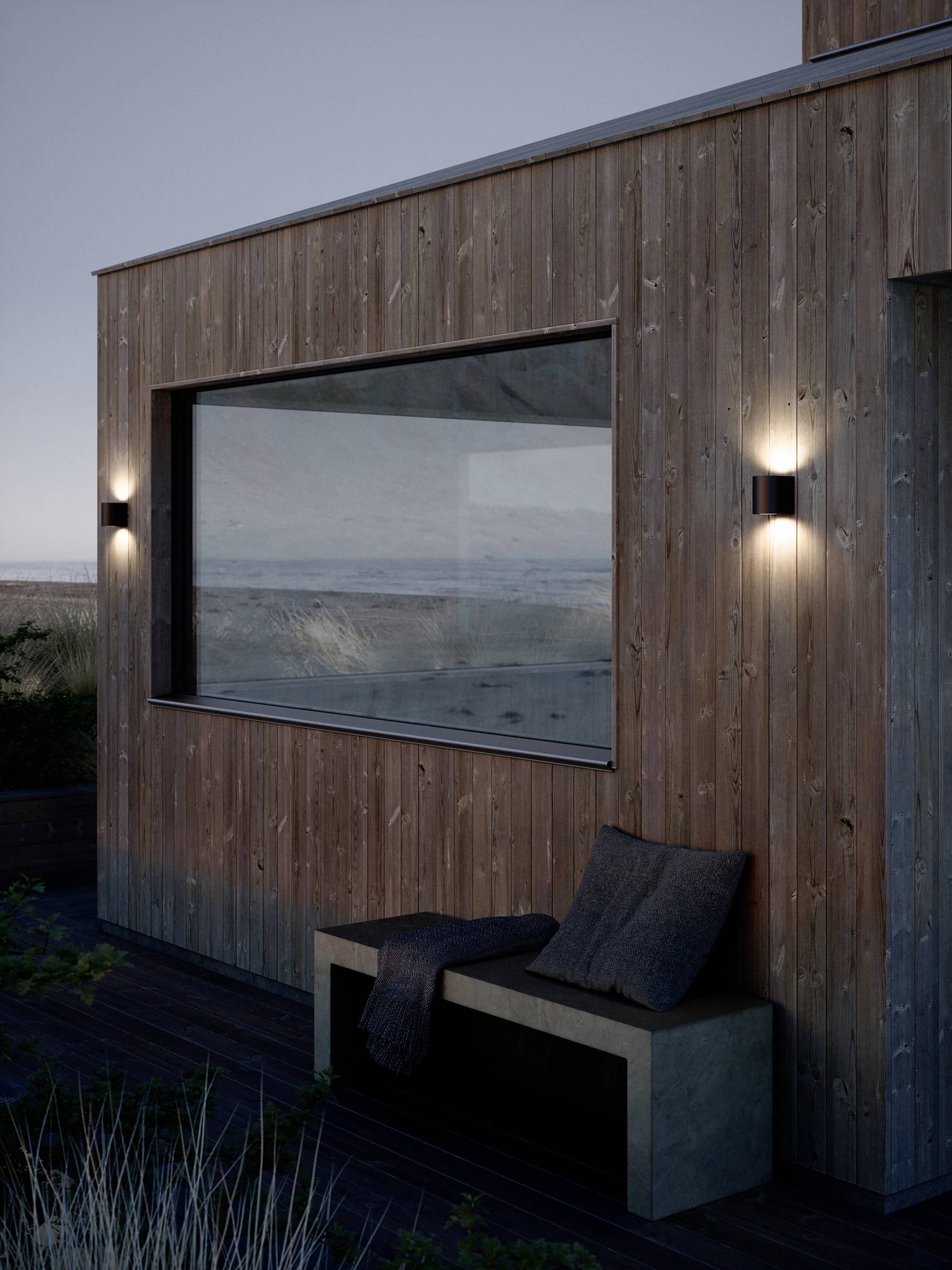Modern houten huis met groot raam, twee wandlampen en een betonnen bank met kussens, omgeven door natuurlijke vegetatie bij schemering.