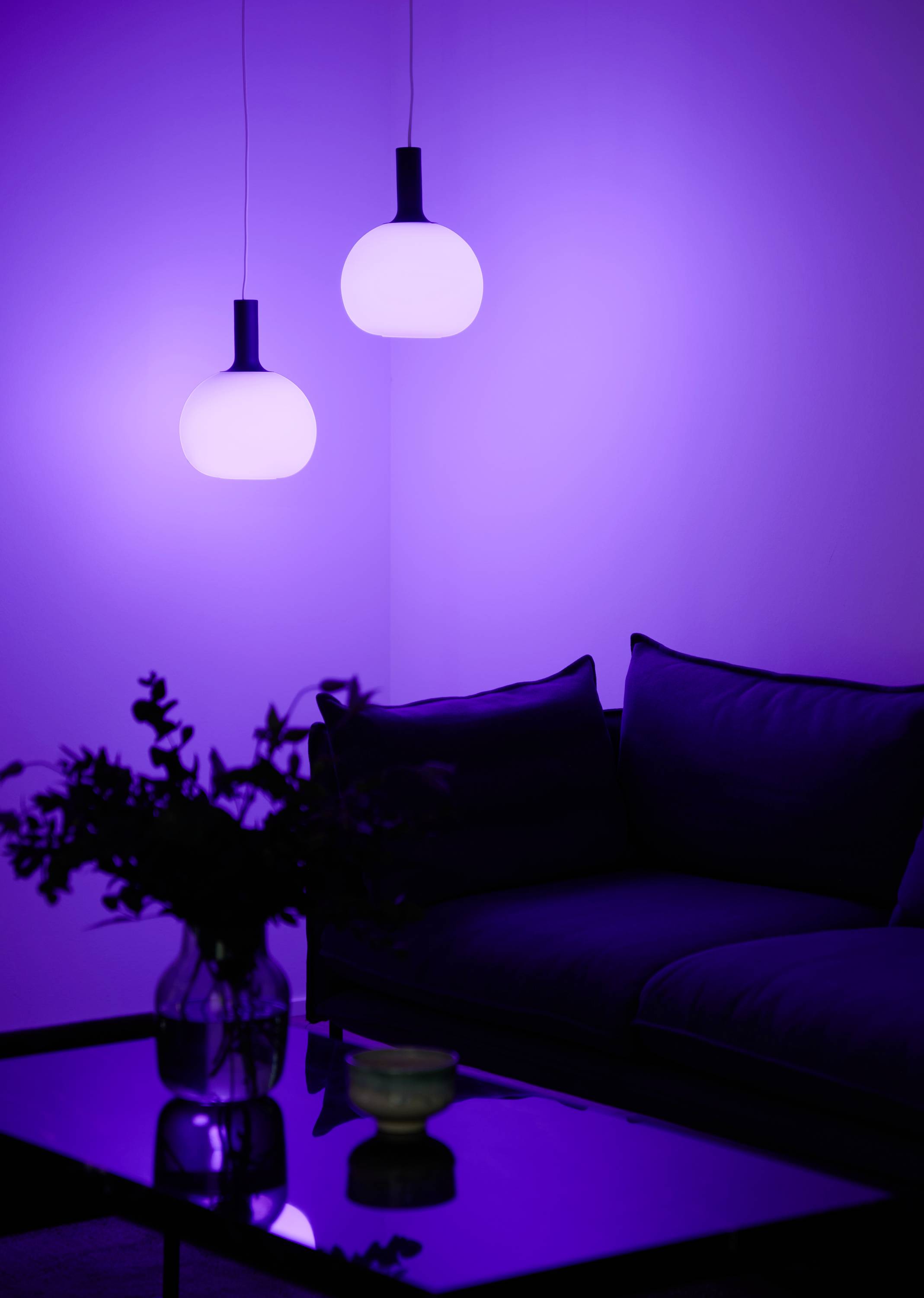 Twee hangende verlichtingsarmaturen in een moderne interieurinrichting die een paarse gloed werpen op een bank en een glazen tafel.