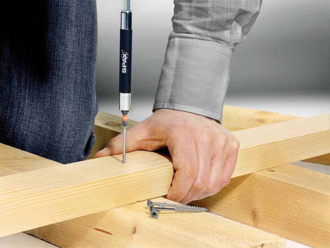 Een hand houdt een houten plank vast terwijl een schroevendraaier een schroef draait die de plank aan een houten constructie bevestigt.