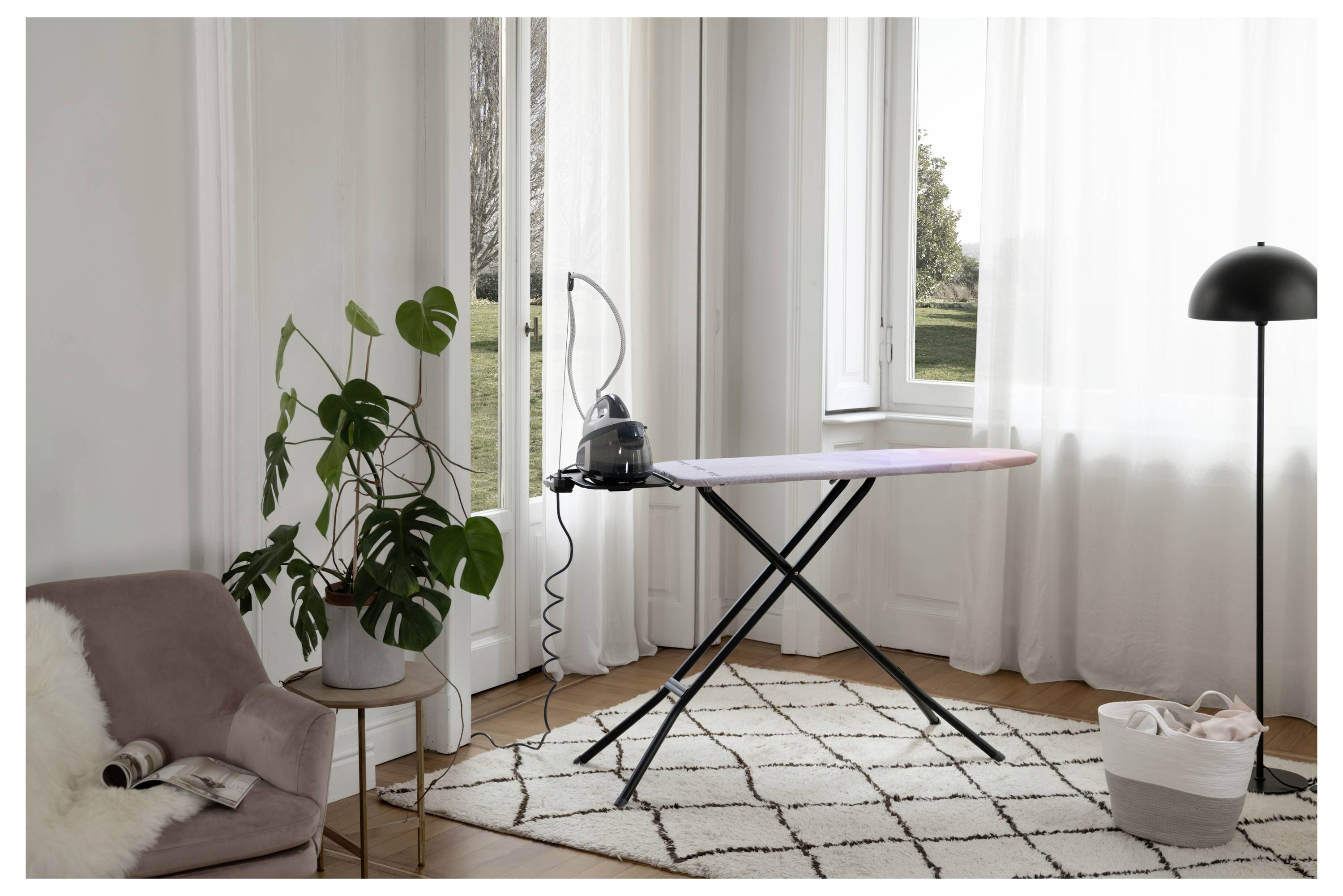 Lichte woonkamer met een compact strijkplank bij het raam, naast een staande lamp, potplant, stoel en wasemand.