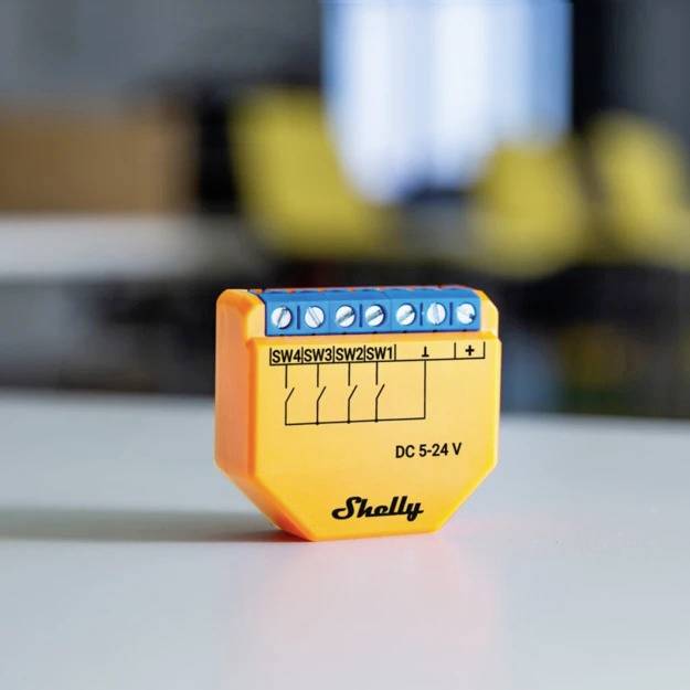 Een klein oranje apparaat met blauwe aansluitingen en het opschrift 'Shelly DC 5-24 V' op een tafel, onscherpe achtergrond.