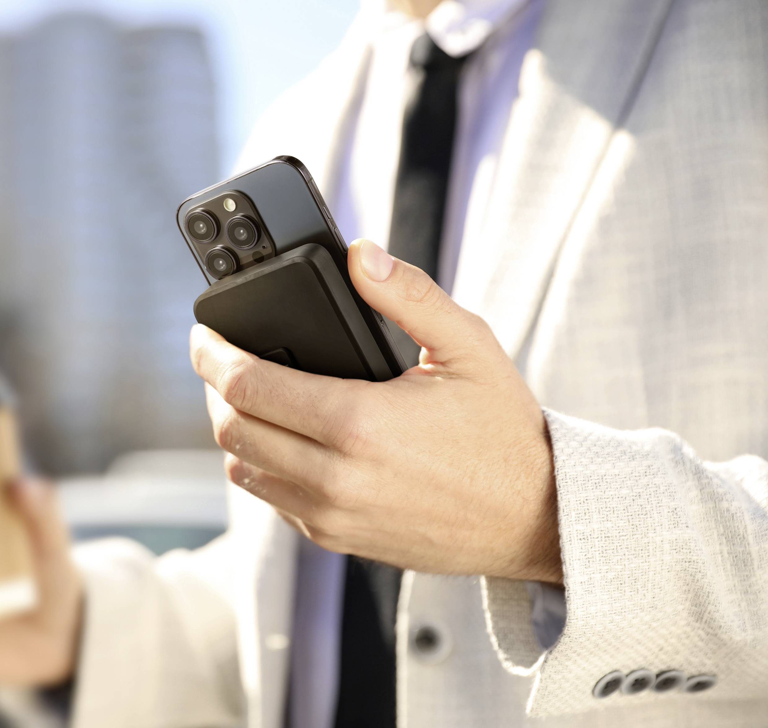 Een man in een licht pak houdt een smartphone en een koffiekopje vast. Op de achtergrond zijn onscherpe gebouwen zichtbaar.